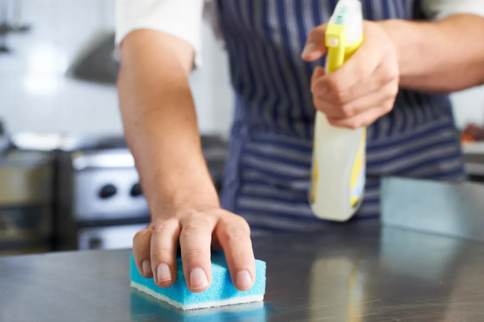 A chef cleaning a metal surface with a blue sponge while holding a spray bottle.