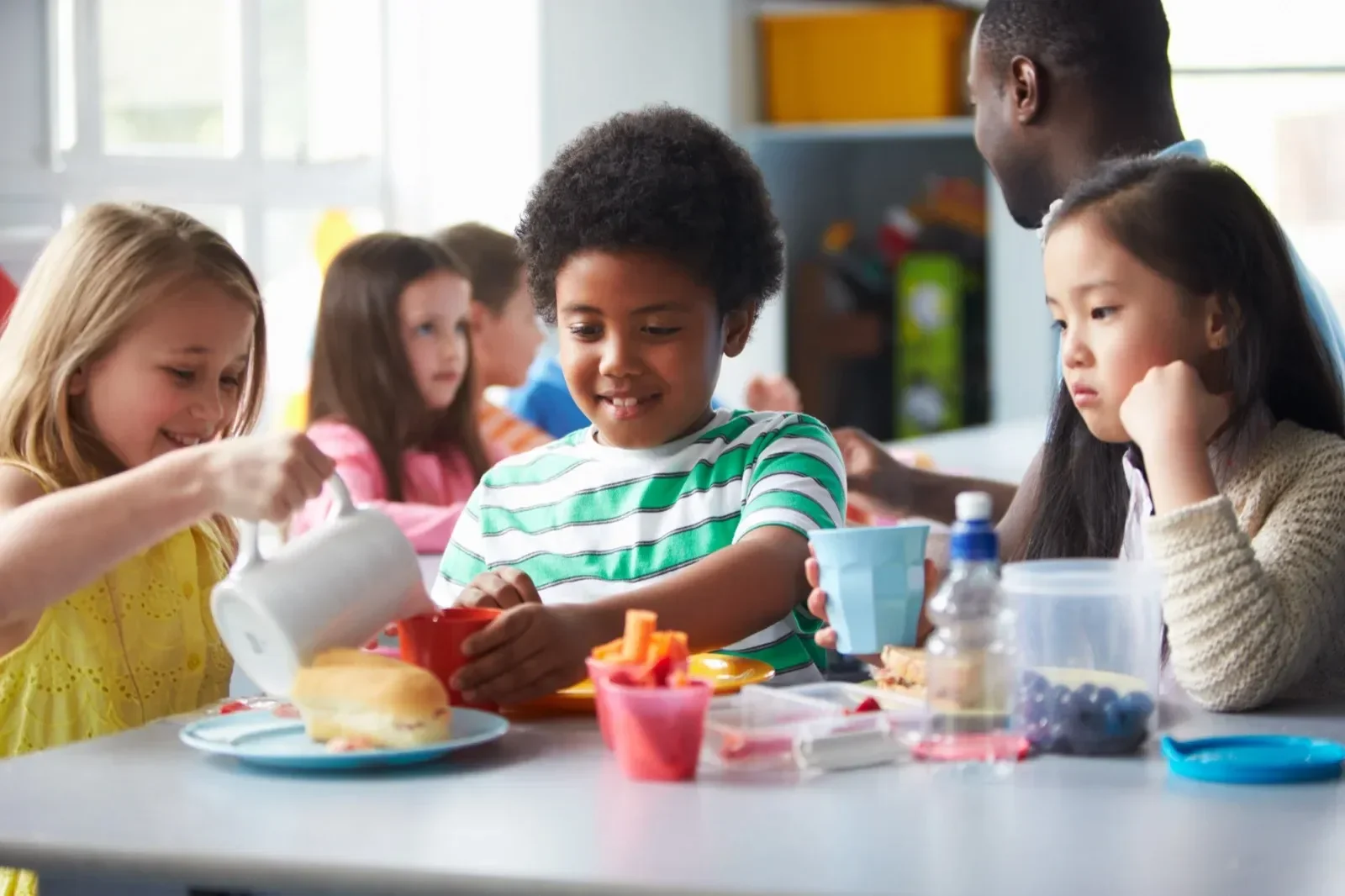 A diverse group of children sitting at a table during lunch, with some pouring juice and others looking at their food.