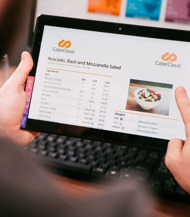 Person holding a tablet displaying a recipe for Avocado, Basil, and Mozzarella Salad from CaterCloud, with ingredients, quantities, and allergens listed, alongside a photo of a bowl of salad.
