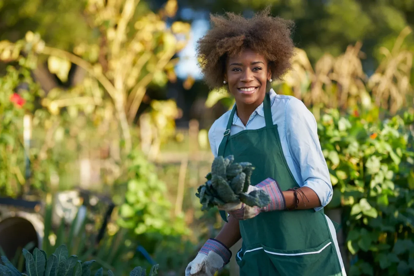A woman in a green apron and gloves is smiling while working in a garden with green plants and trees in the background.