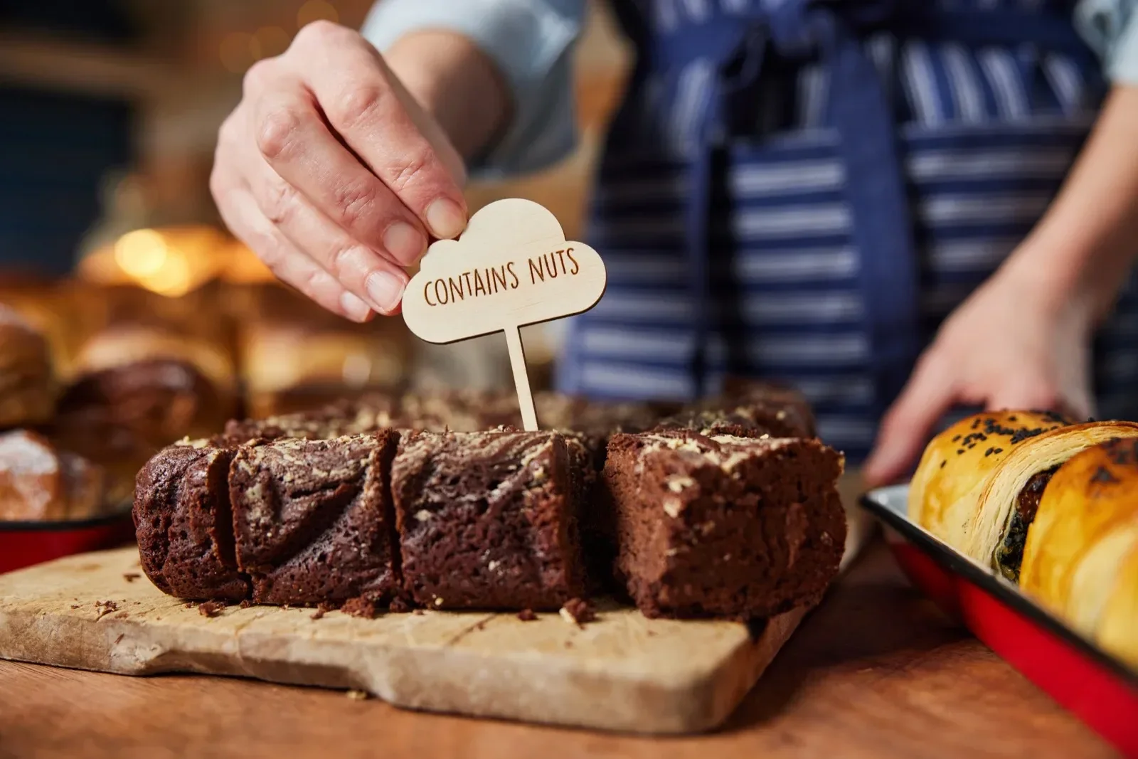 A person is holding a sign that says 'Contains Nuts' above slices of chocolate brownies on a wooden cutting board in a bakery or cafe.