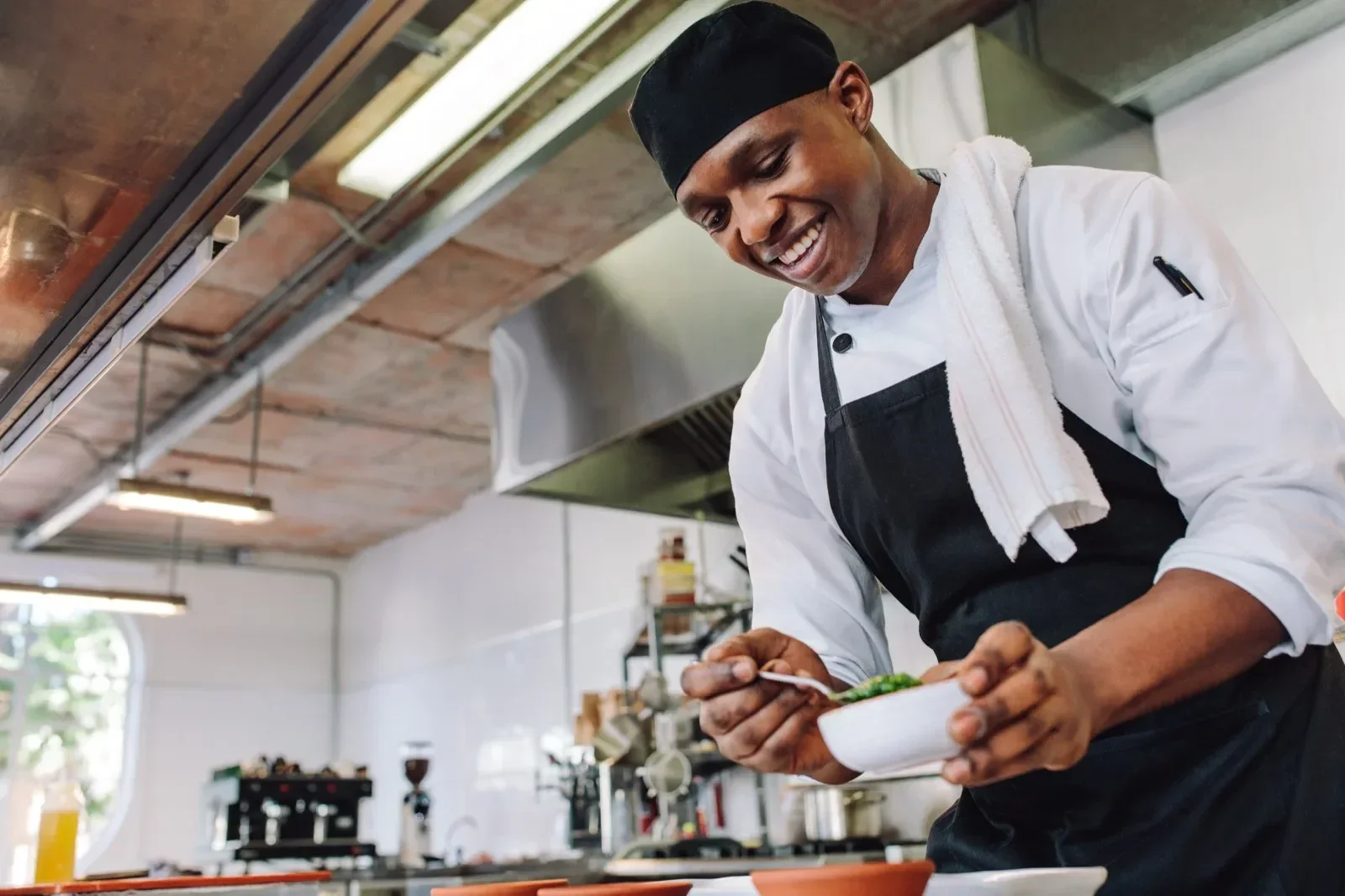 A smiling male chef in a black hat and apron preparing food in a modern, industrial-style kitchen.