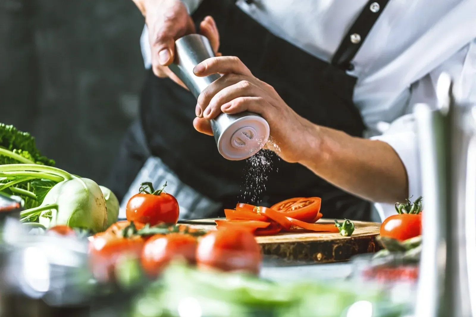 Person seasoning cut tomatoes and slicing them on a wooden cutting board, with fresh vegetables such as broccoli and radishes on the counter.