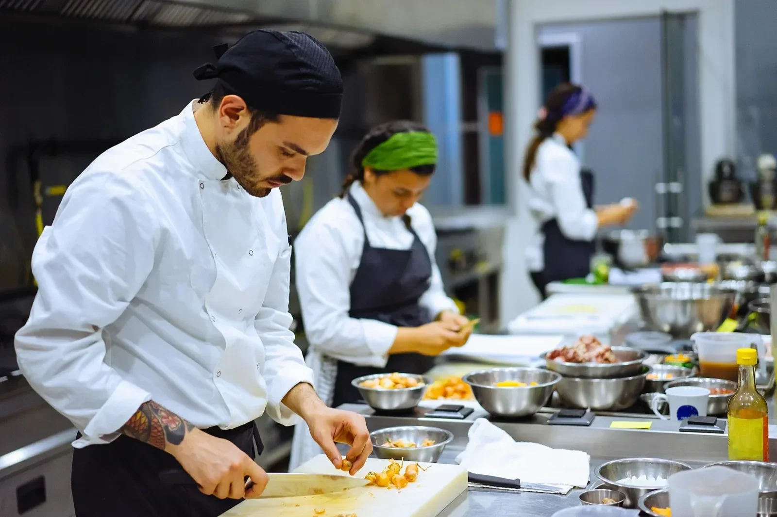 Three chefs in a professional kitchen preparing food. The chef in the foreground is chopping ingredients on a cutting board, with various bowls and kitchen utensils around.