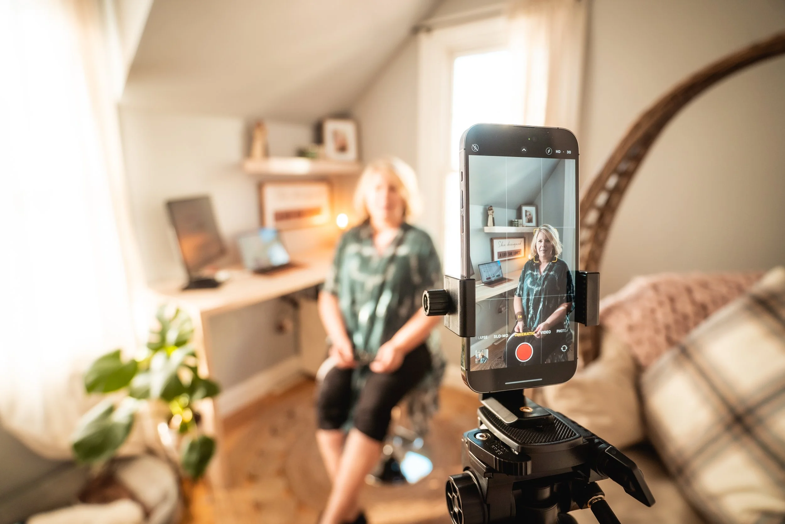 A woman sitting on a stool is being filmed with a smartphone mounted on a tripod in a well-lit room with a sofa, a desk with a computer, and decorative items on a shelf in the background.