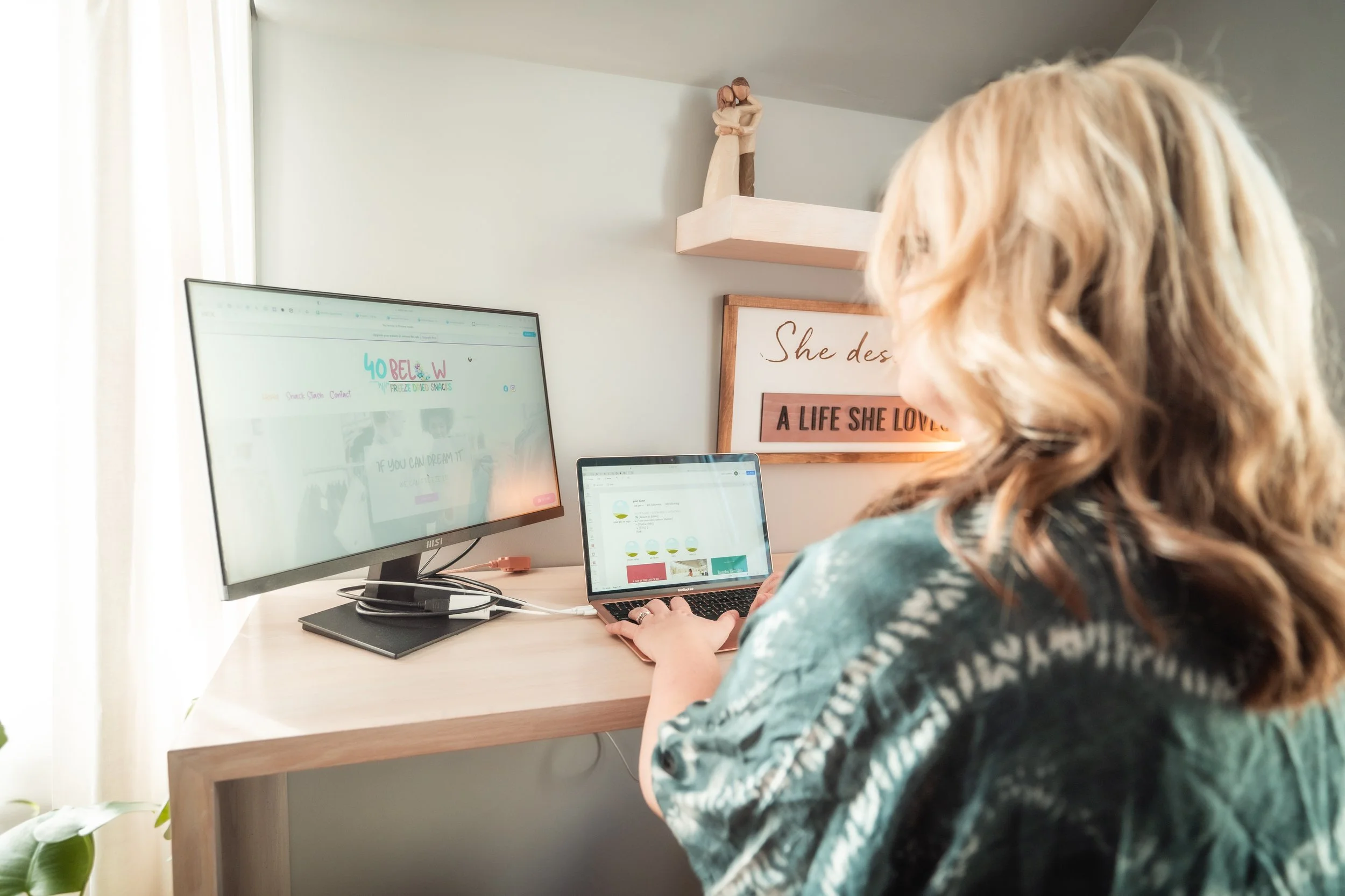 A woman with wavy blonde hair working at a desk with a large monitor and a laptop, with a wall decorated with a sign reading, "She des... A LIFE SHE LOVED."