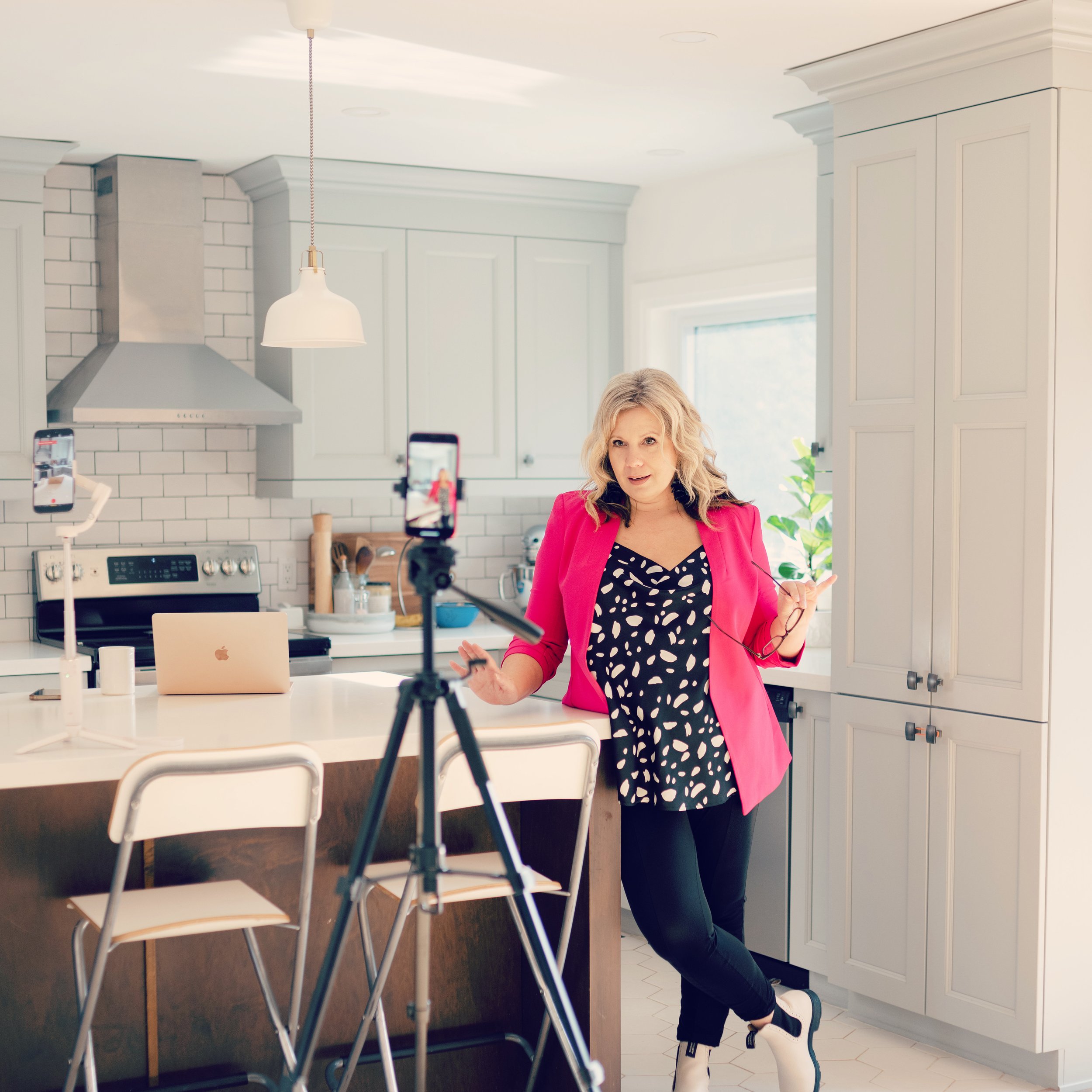 A woman in a pink blazer and black dress is filming a video in a kitchen with white cabinets and a white tiled backsplash, standing by a white kitchen island with a laptop and camera setup.