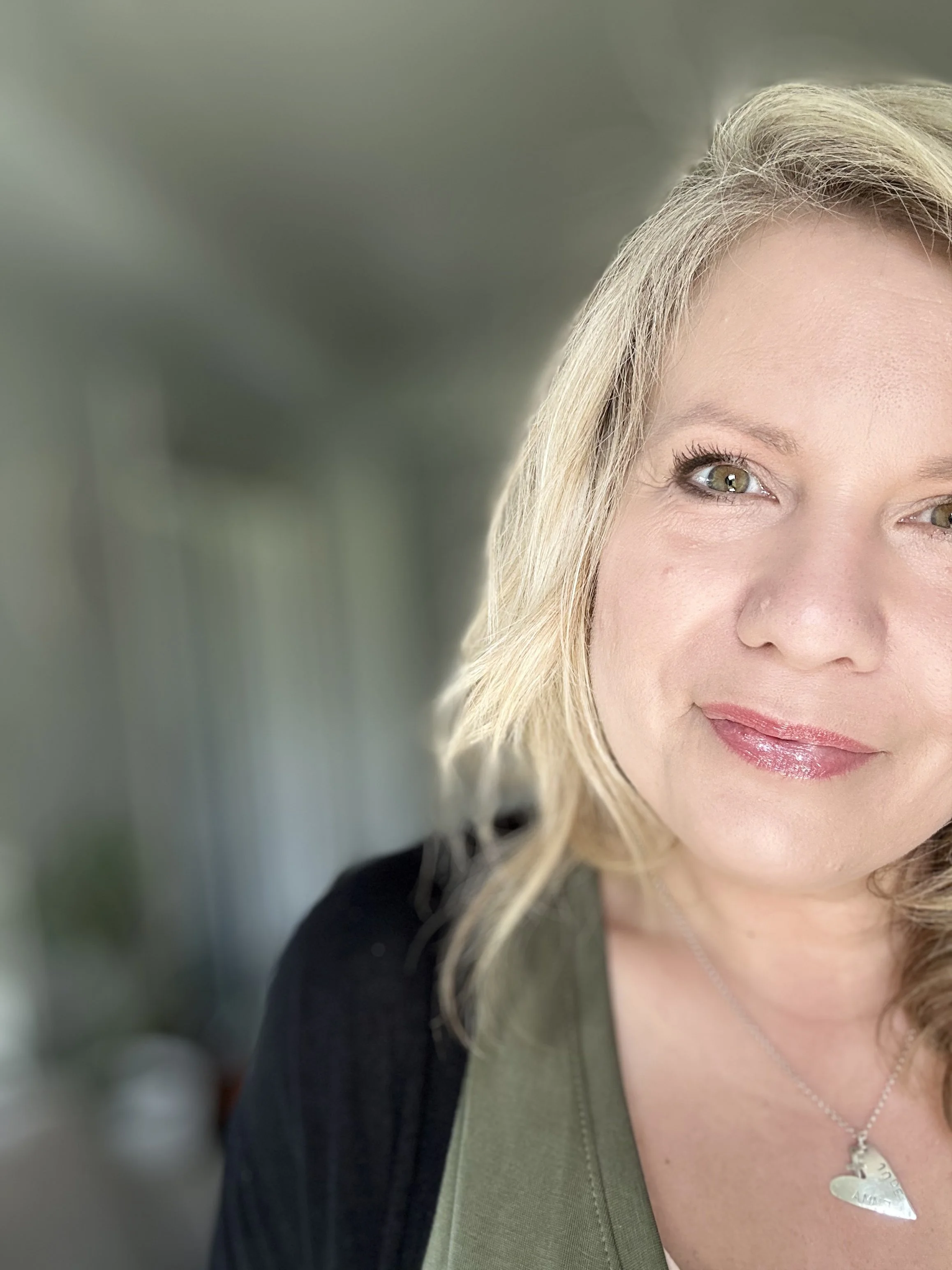 Close-up of a woman with blonde hair, wearing a green top, black jacket, and a silver necklace, smiling outdoors with a blurred background.