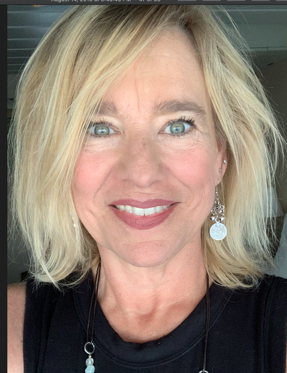Close-up of a woman's smiling face with blonde hair, blue eyes, pink lipstick, wearing earrings and a black top.