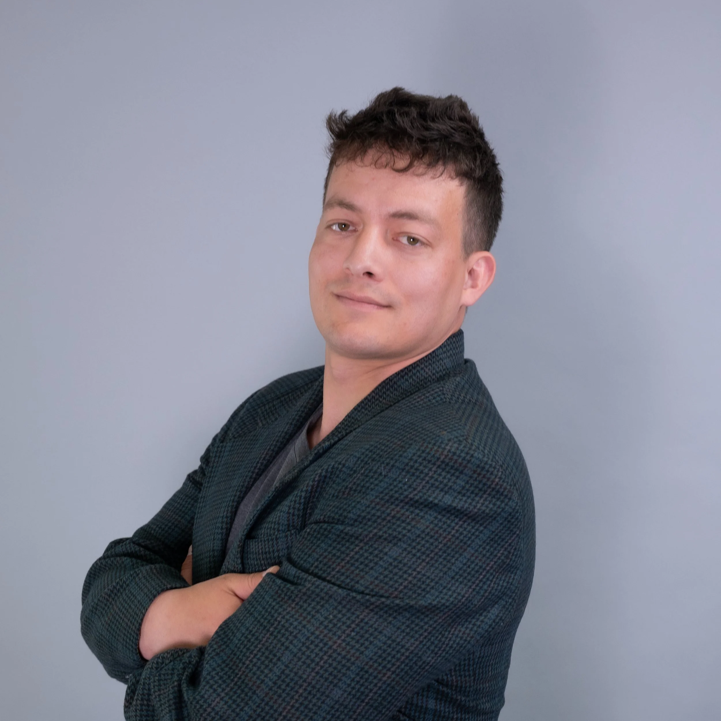 Portrait of a young man with dark hair wearing a dark blazer, standing against a plain light gray background, with arms crossed and a slight smile.