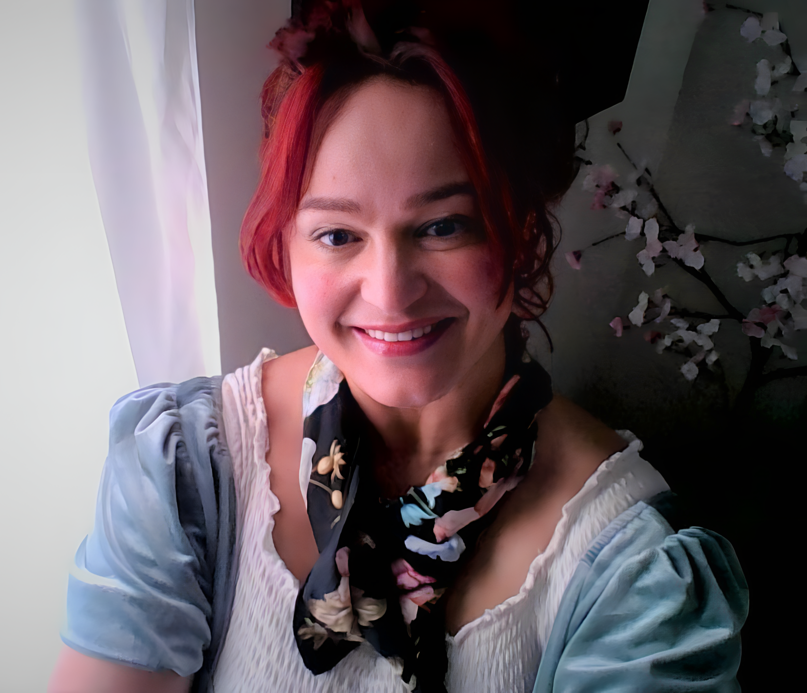 A smiling woman with short red hair, wearing a light-colored top and a dark floral scarf, standing indoors near a white curtain and a background of flowers.