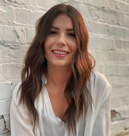 Young woman with long, wavy brown hair smiling, wearing a white top, standing in front of a white brick wall.