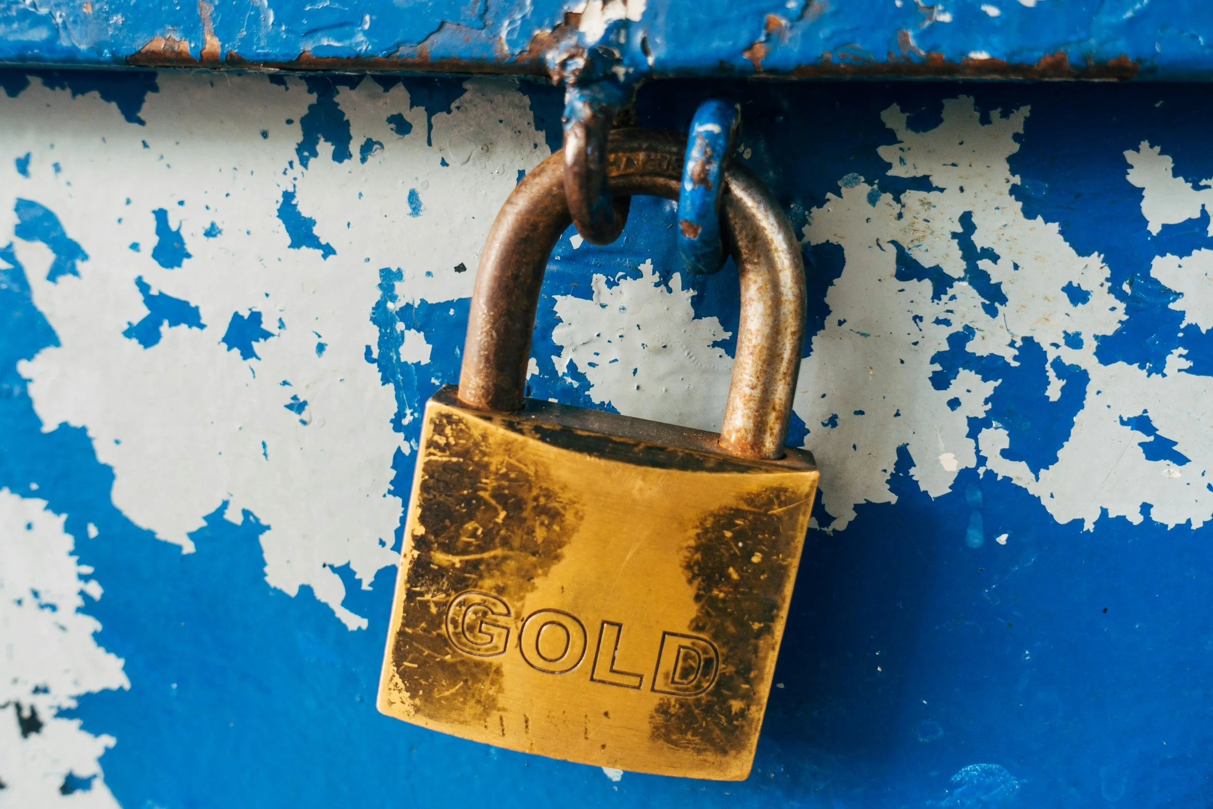 Close-up of a gold padlock with the word 'GOLD' engraved on it, attached to a rusty blue latch on a weathered, chipped blue and white painted surface.