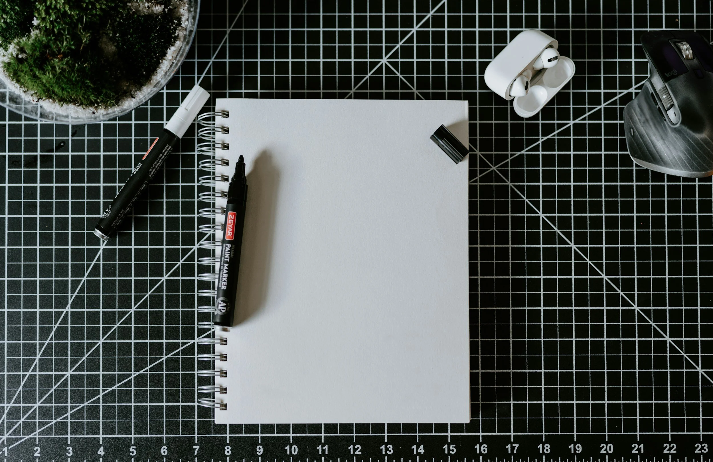 A workspace with a spiral-bound blank notebook, black marker, black marker cap, white wireless earbuds, wireless mouse, and a plant in a glass container on a black cutting mat with white grid lines.
