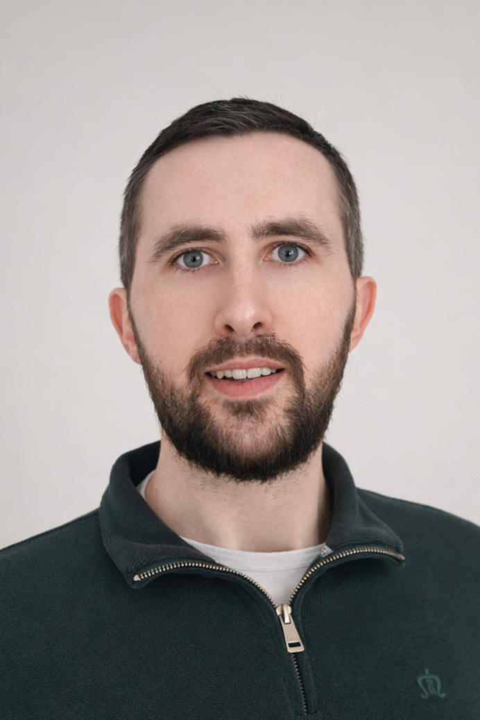 A man with short brown hair and a beard, wearing a dark zip-up jacket, looking at the camera with a neutral expression against a plain light background.