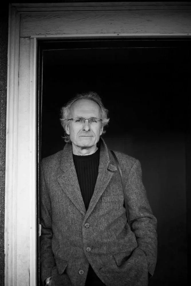 An older man with glasses, gray hair, and a serious expression, standing in a doorway, wearing a dark turtleneck and a tweed blazer.