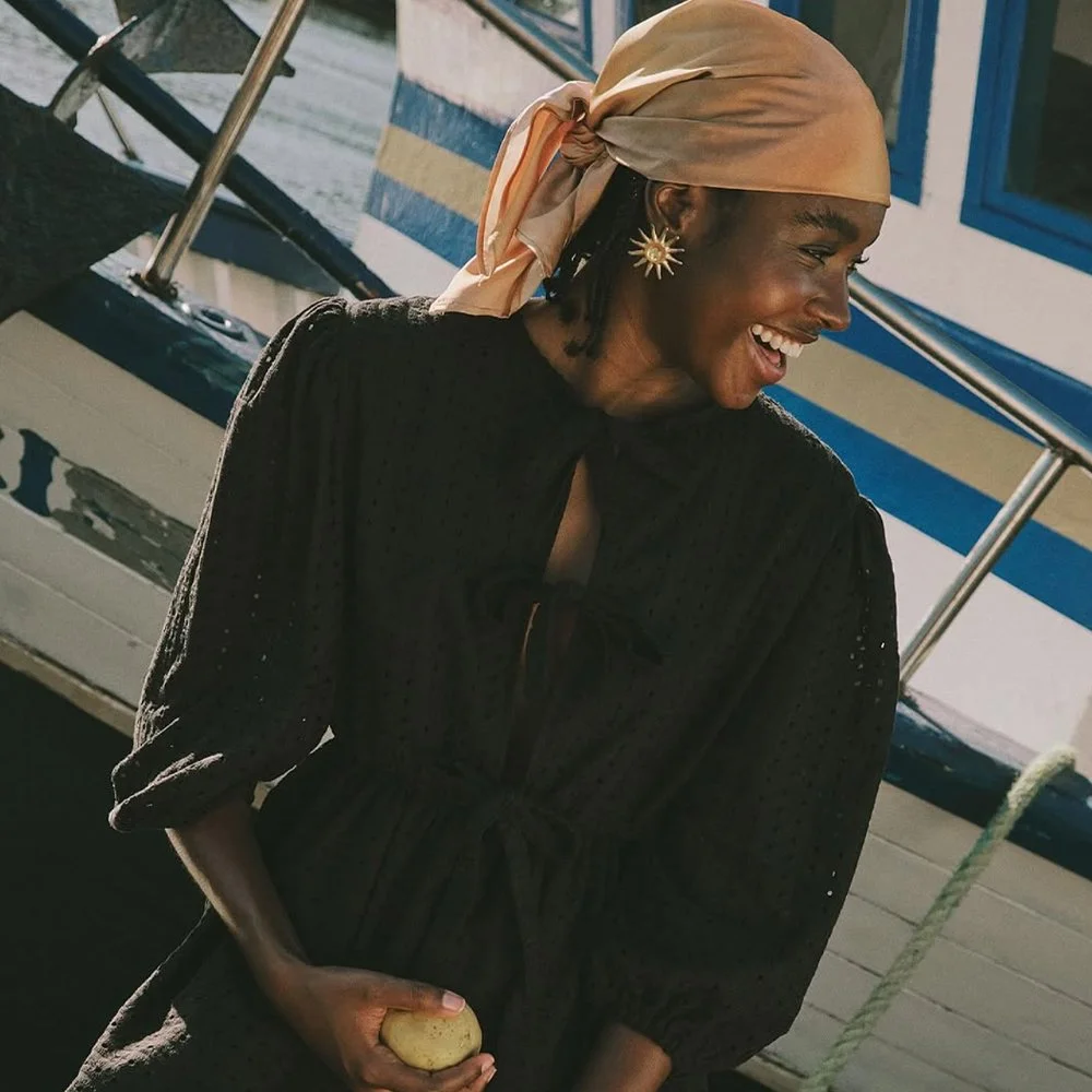 Mulher sorrindo, usando lenço bege na cabeça, brincos de sol e vestido preto, ao lado de um barco.