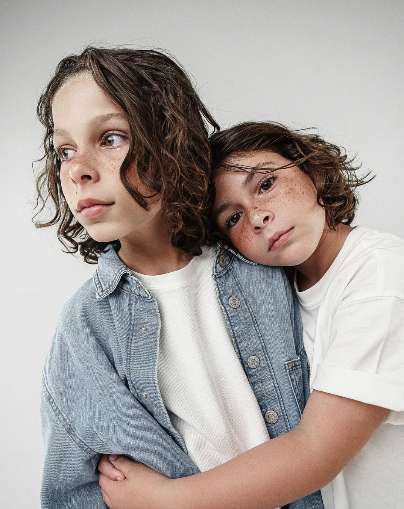 Dois meninos com cabelo cacheado, olhos claros e sardas estão abraçados, um usando jaqueta jeans e o outro camiseta branca, contra fundo branco.