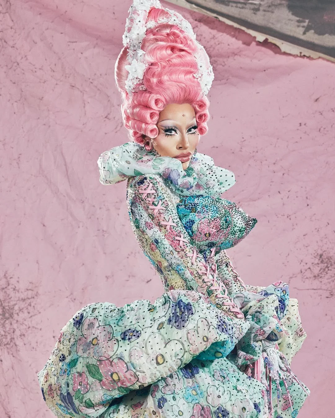 Pessoa com maquiagem elaborada, cabelo rosa em estilo teatral, vestindo roupa com detalhes florais, lãs e decoração colorida, em fundo rosa.