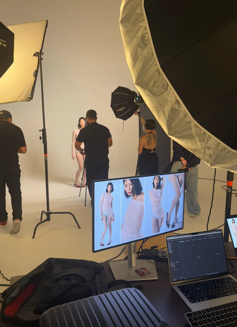 Sessão de fotografia de moda em um estúdio com várias luzes e equipe ao redor, enquanto uma modelo posa com roupas claras e salto alto, com uma tela exibindo as fotos tiradas.