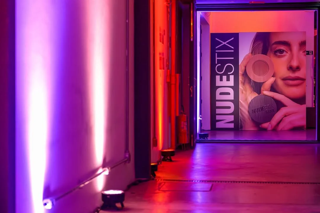 Corredor com iluminação neon roxa, pink e vermelha, com uma parede de espelho na extremidade esquerda e um anúncio publicitário de maquiagem na extremidade direita, exibindo uma mulher segurando dois produtos de maquiagem