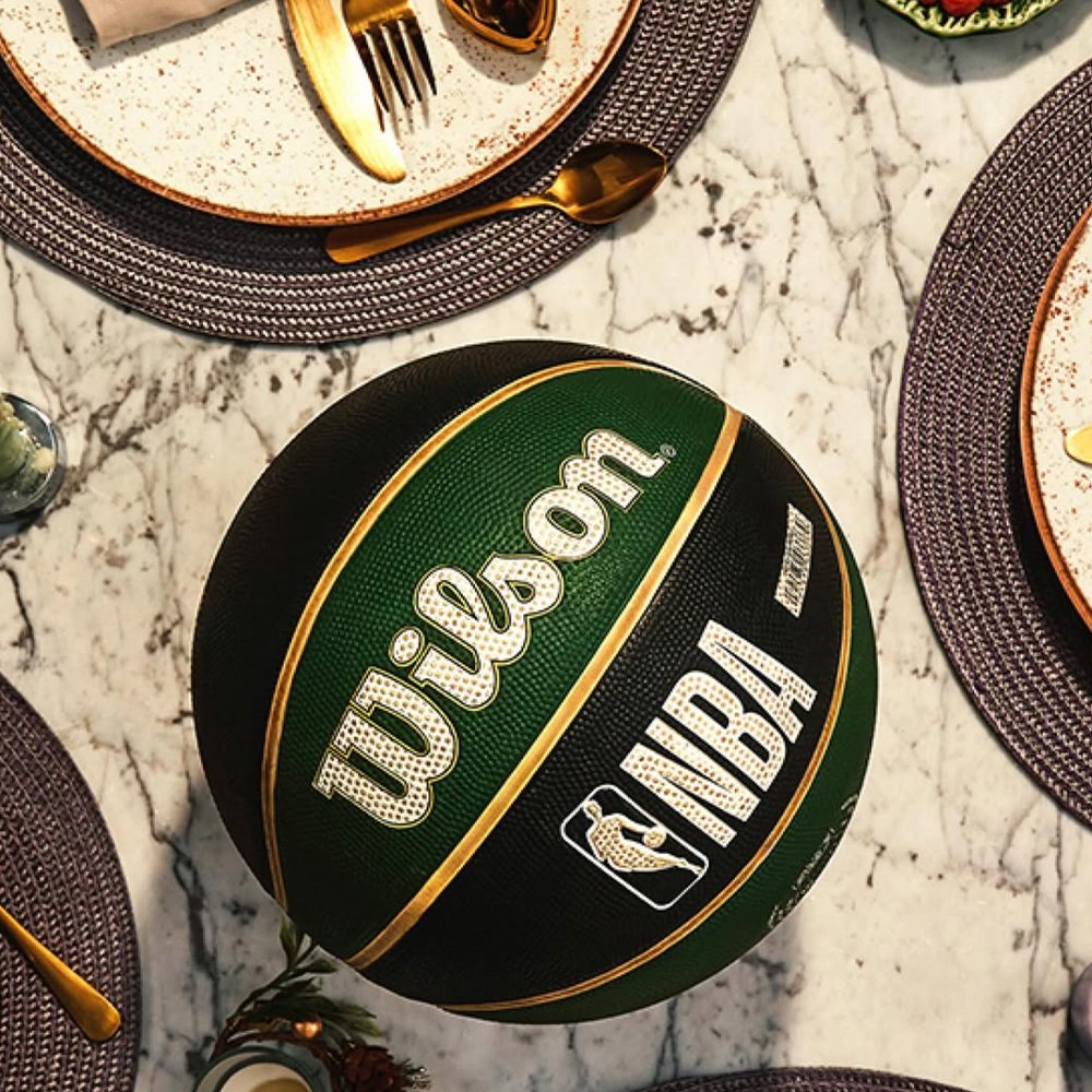 Bola de basquete verde e preta com o logotipo da NBA sobre uma mesa de mármore, cercada por pratos decorativos, talheres dourados e copos.