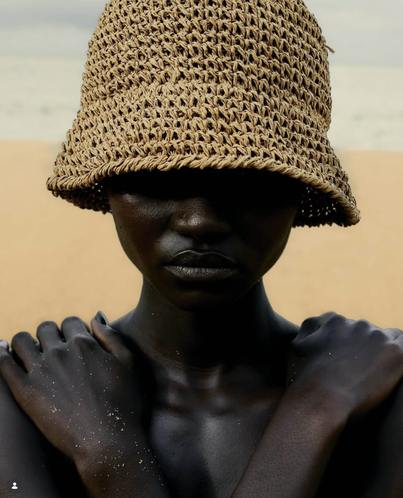 Mulher usando um chapéu de palha, cobrindo parcialmente seu rosto, com uma praia ao fundo.