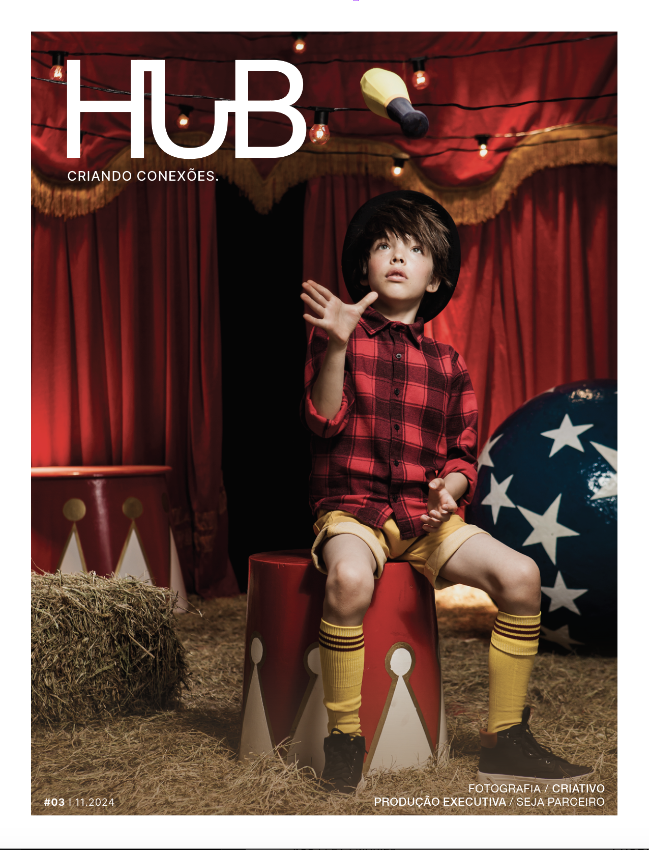 Capa de revista com menino sentado em palco de circo, usando boné preto, camisa vermelha xadrez, shorts amarelos e meia amarela com riscas pretas, rodeado por decoração de circo e lona vermelha.