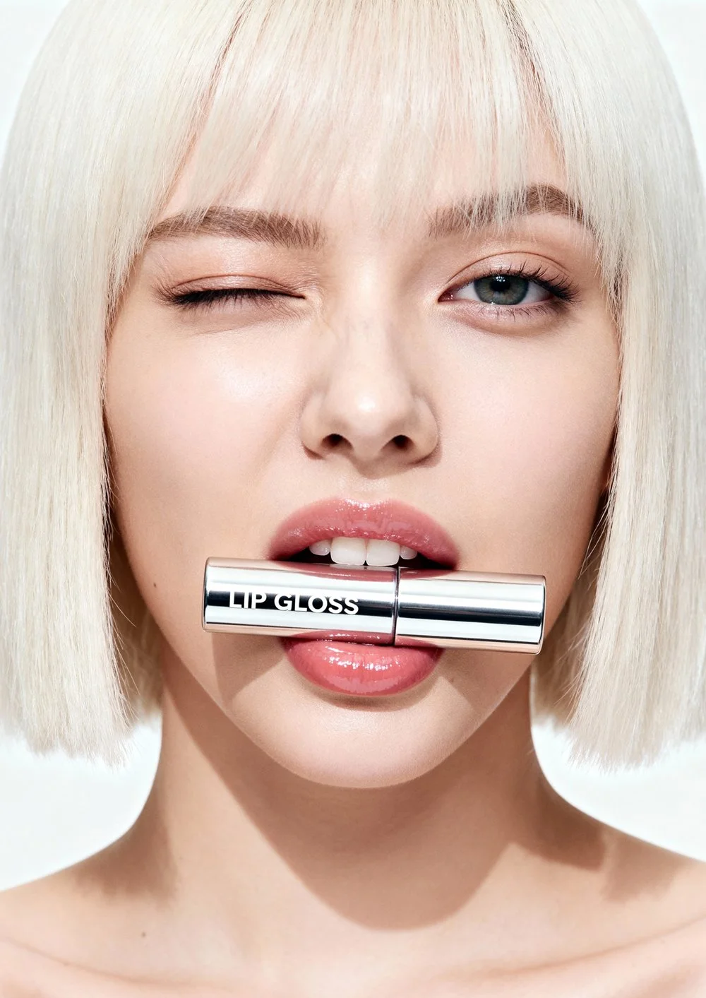 Mulher de cabelo platinado, uma olhando para a câmera com olho fechado e boca levemente entreaberta, segurando um batom labial prateado com a inscrição "LIP GLOSS" na boca.