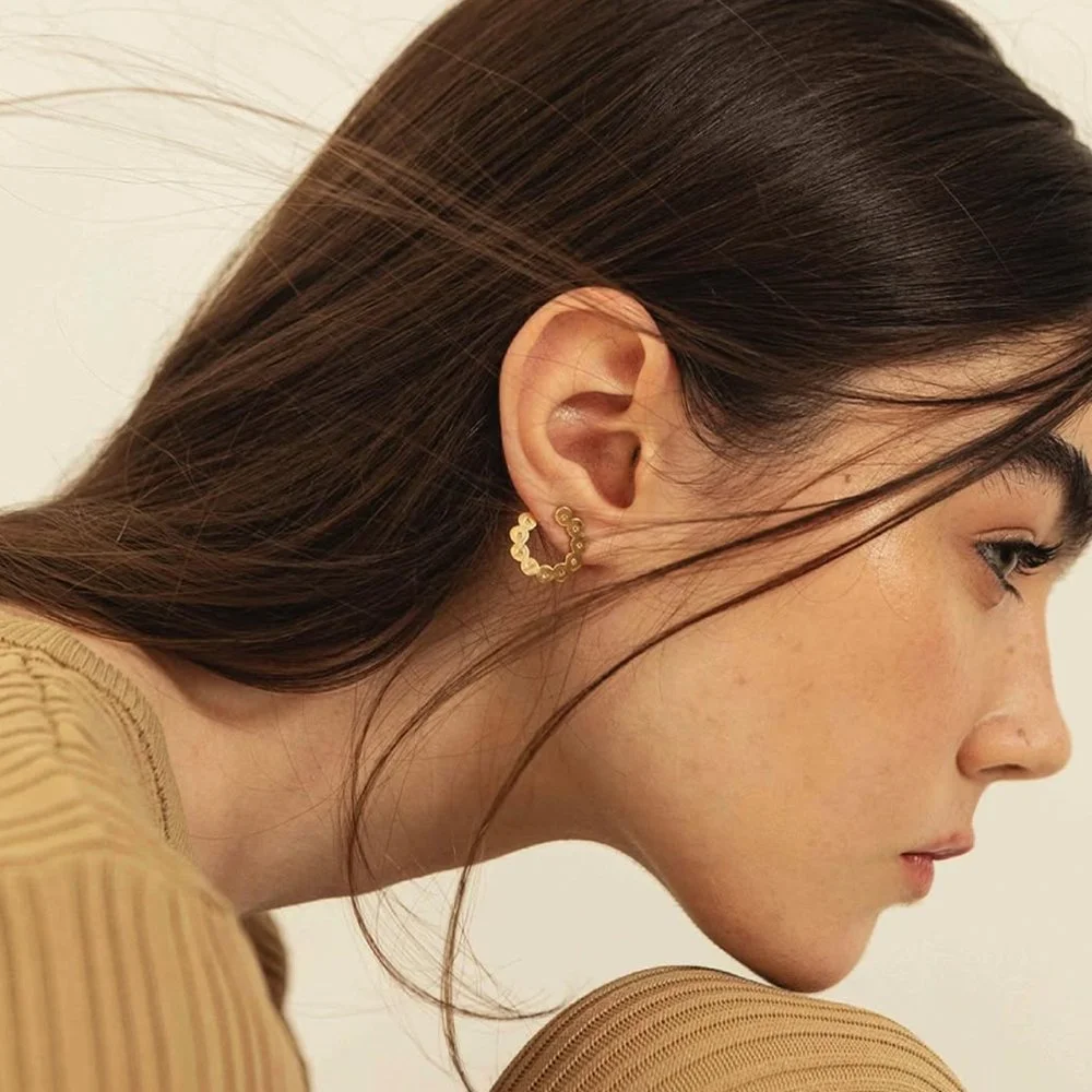 Close-up de perfil de uma mulher jovem com cabelo castanho escuro liso e pequeno, usando brincos dourados em forma de círculo com detalhes pequenos.