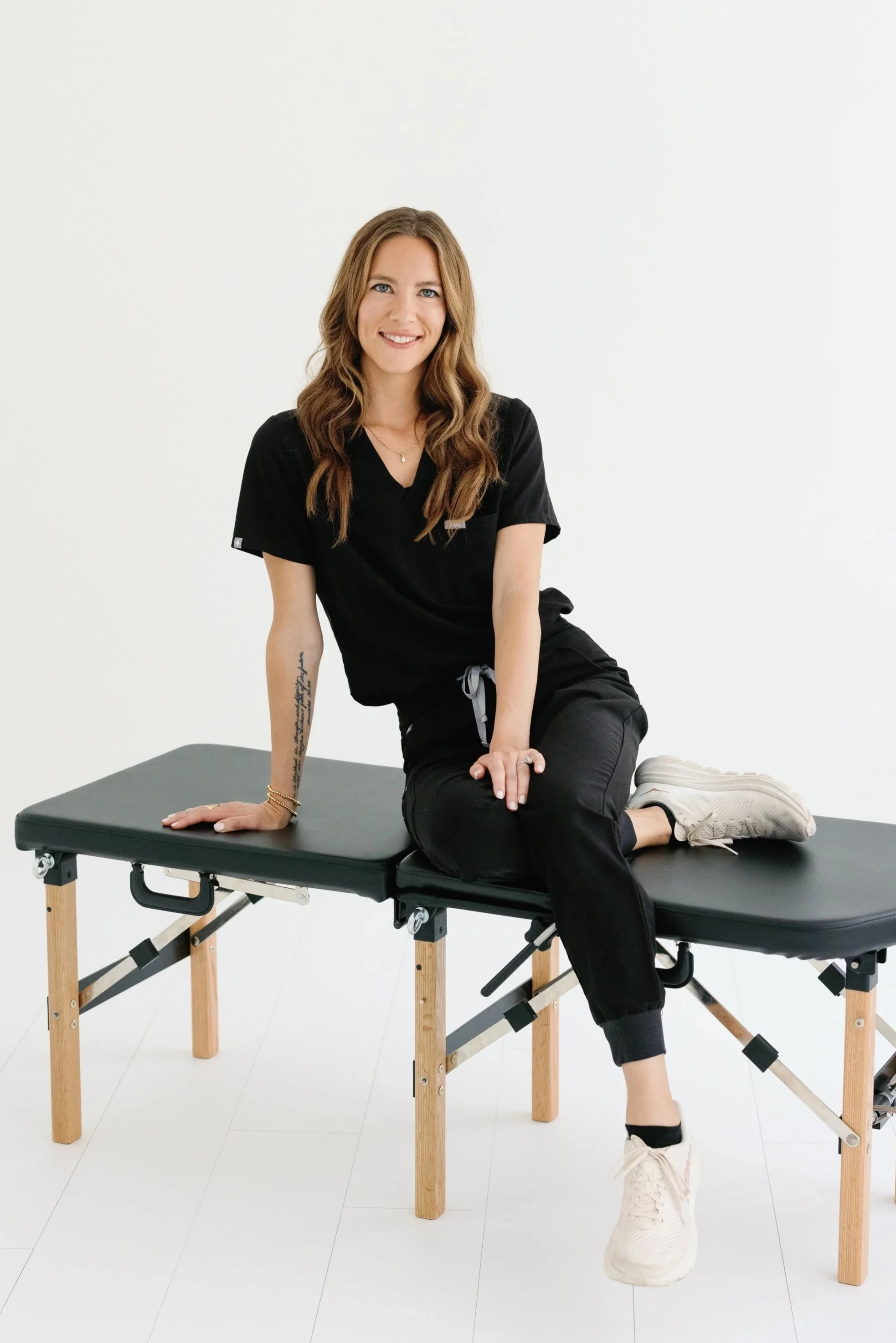 Woman sitting on a black chiropractic table with a white background, wearing black scrubs and white sneakers, smiling at the camera.