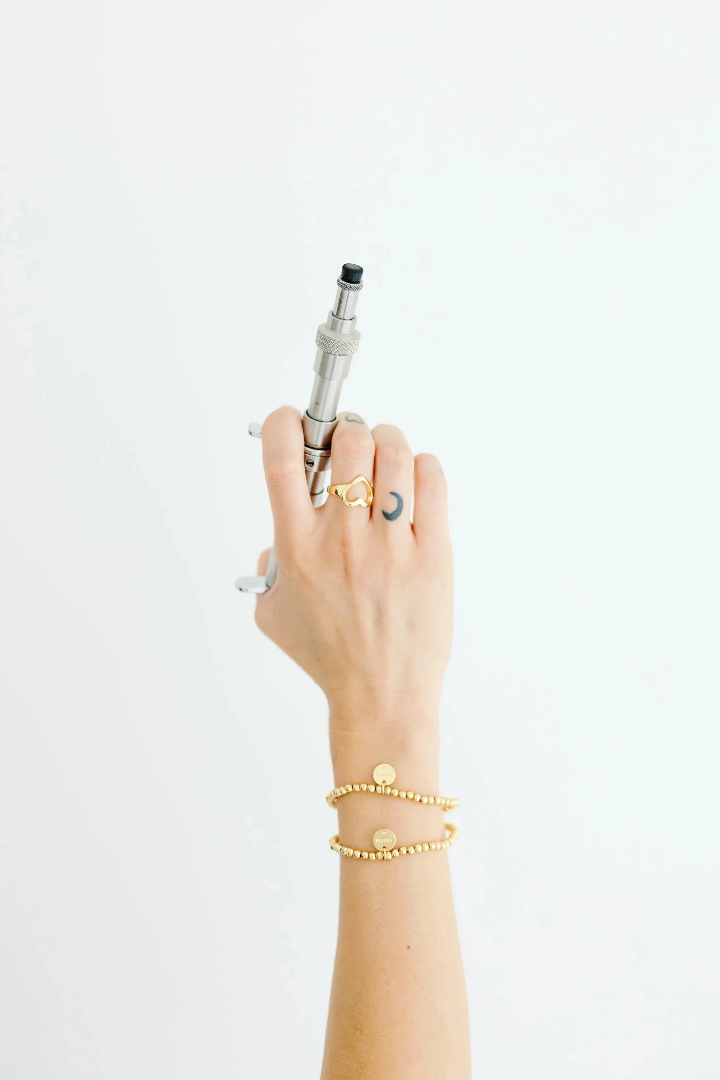 Hand holding a chiropractic adjusting tool/device against a white background, wearing jewelry including a ring, two bracelets, and a tattoo on the index finger.