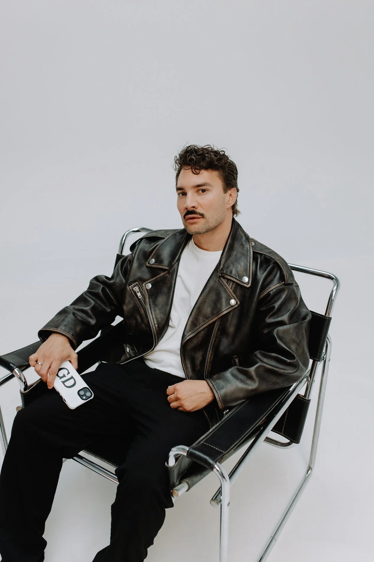 Man zit op een zwarte stoel met metalen frame, draagt een zwarte leren jas en witte T-shirt, houdt een smartphone met 'GD' op de achterkant, heeft donker krullend haar en een snor.