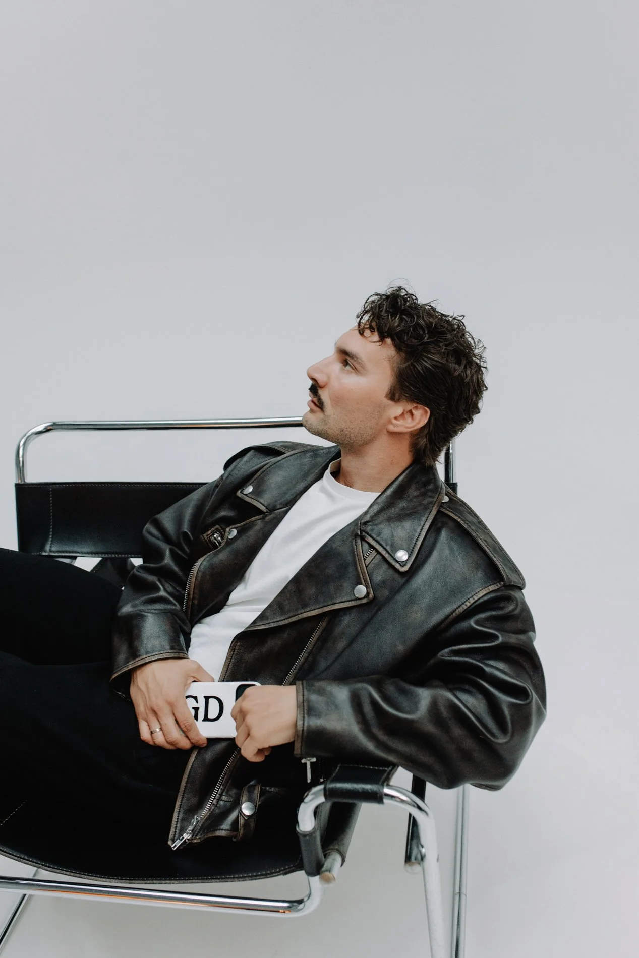 Man zit op een zwarte stoel, draagt een zwarte leren jas en een wit T-shirt, houdt een stuk papier vast en kijkt naar boven, tegen een witte muur.