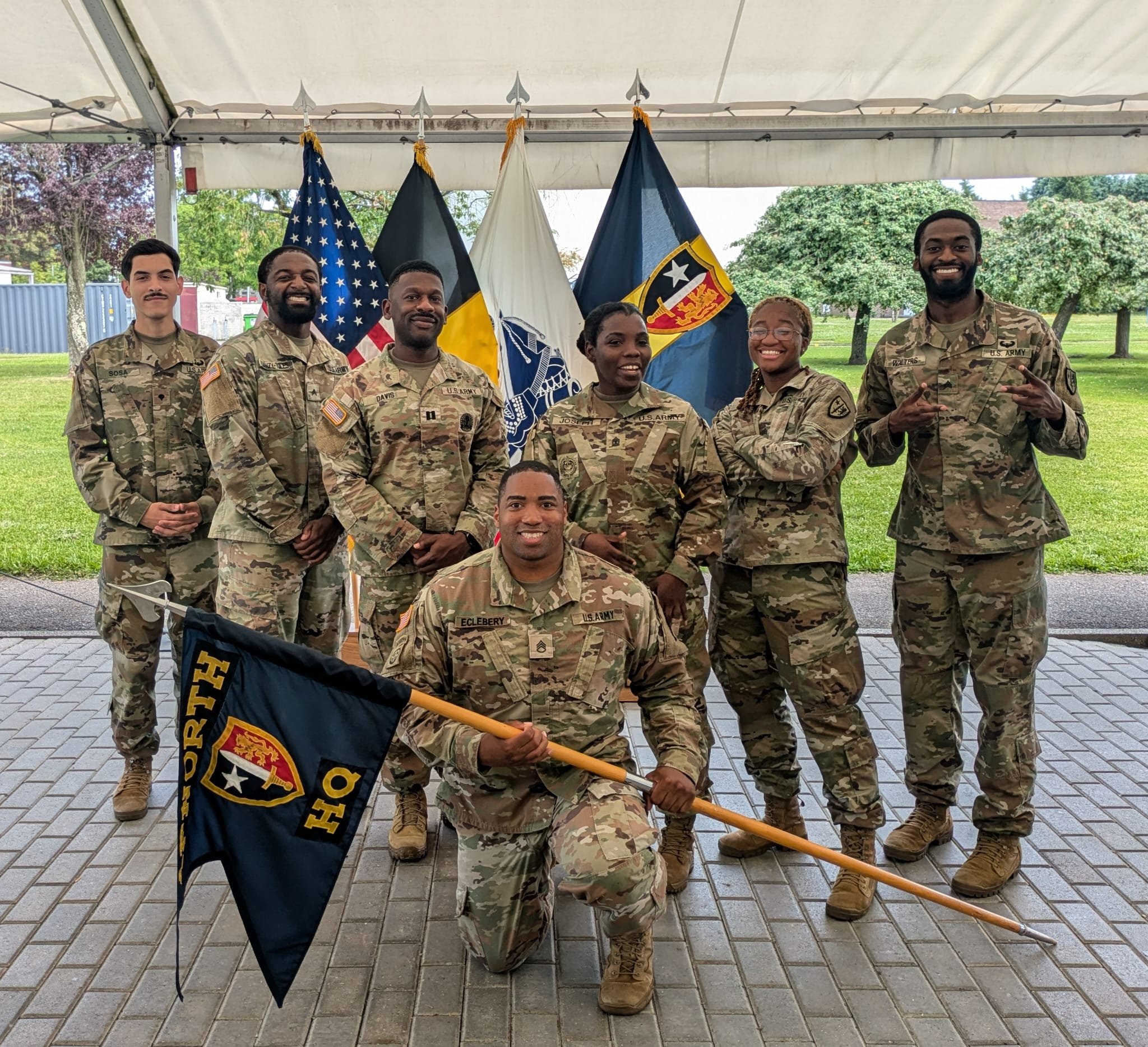 Group of eight Army soldiers posing outdoors under a tent with flags in the background, one soldier kneeling holding a flag, in camouflage uniforms.