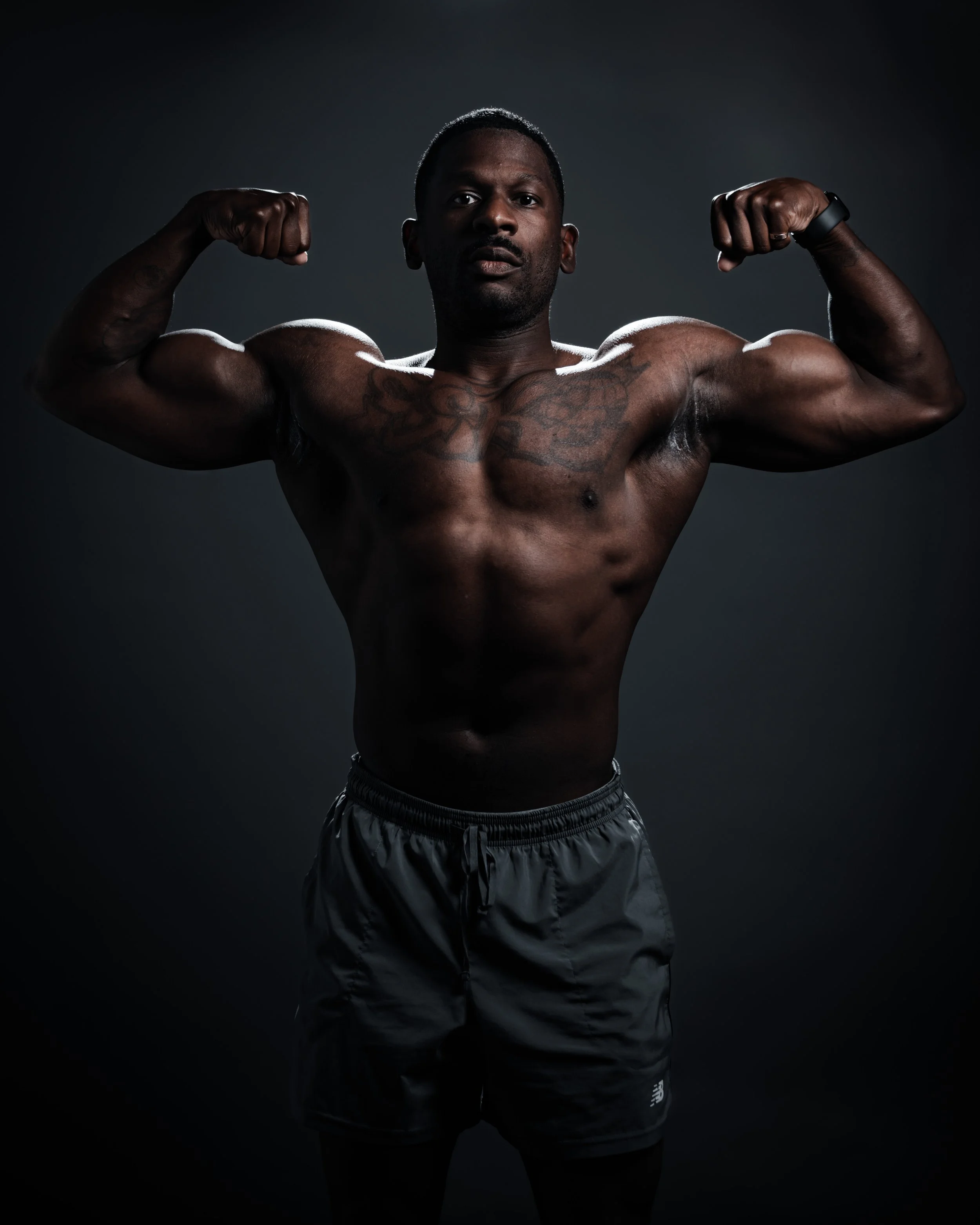 A muscular man with dark skin flexing his biceps in a dark studio setting.