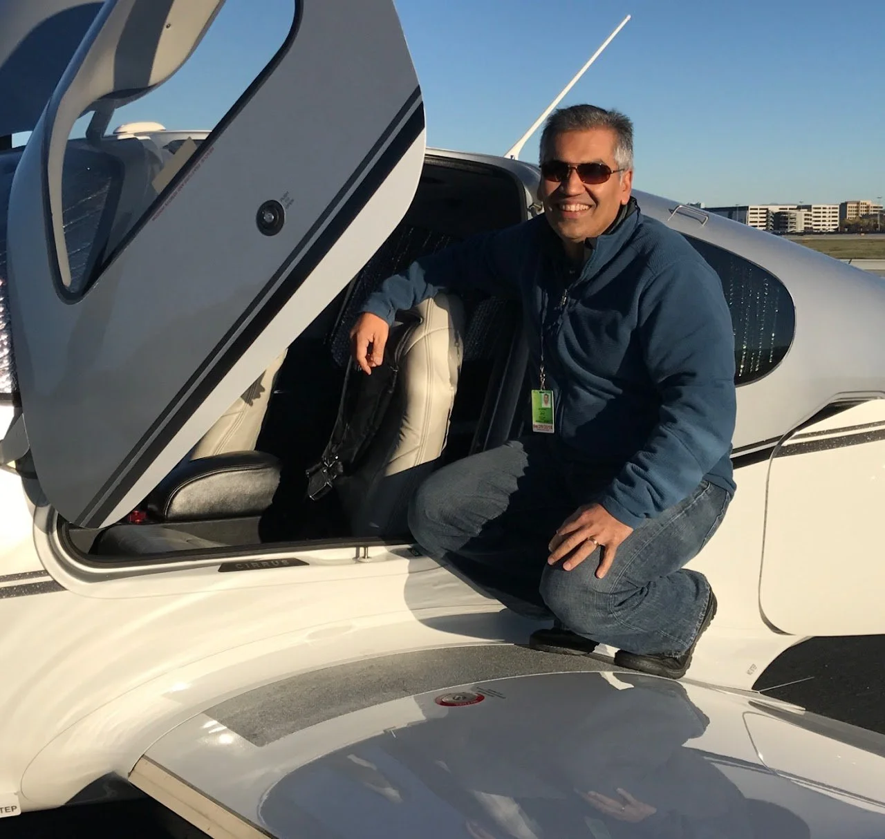 A man in sunglasses, a blue jacket, and jeans is crouching next to a small white aircraft with its cockpit canopy open. The background shows a clear blue sky and distant buildings.