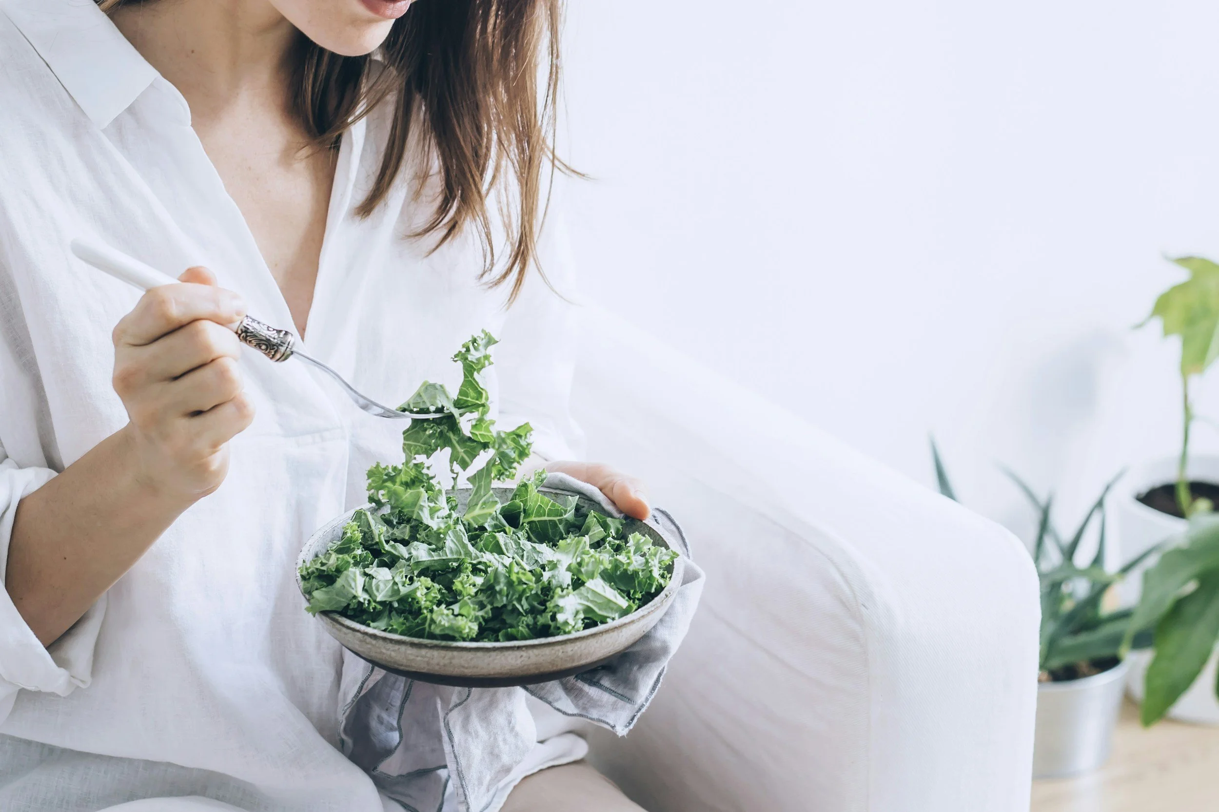 A woman in a white shirt sitting on a white couch, holding a bowl of salad and a fork, with blurred green potted plants in the background.