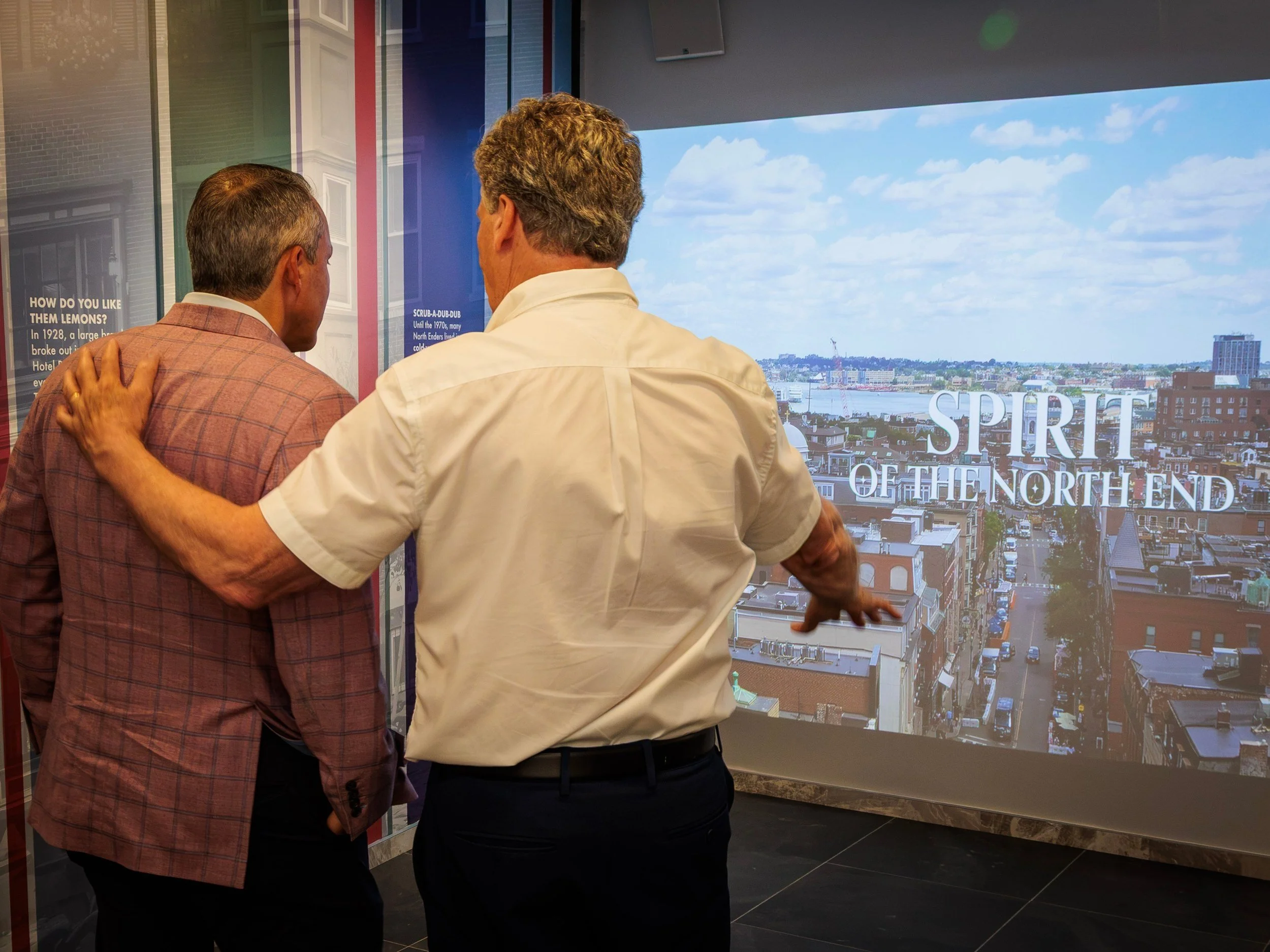 Spirit of the North End playing on the full-height projection wall, opening on a rooftop view of the neighborhood and harbor.