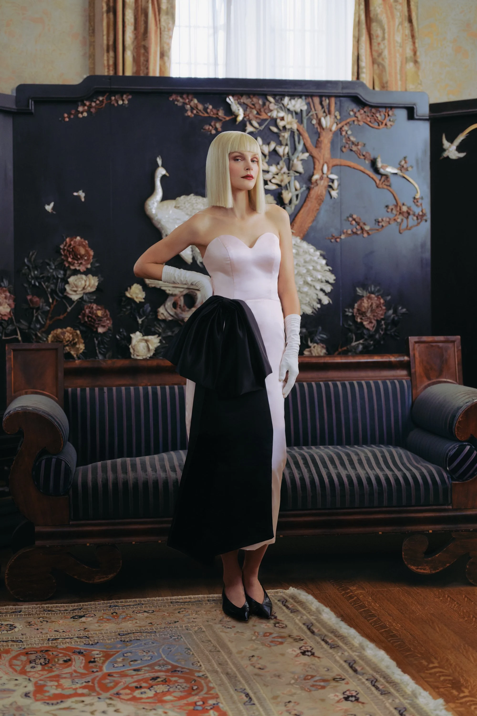 A woman in a strapless light pink dress with black accents stands in front of an ornate wooden and fabric sofa with a floral and bird-themed background behind her. She is wearing white gloves and black high heels.