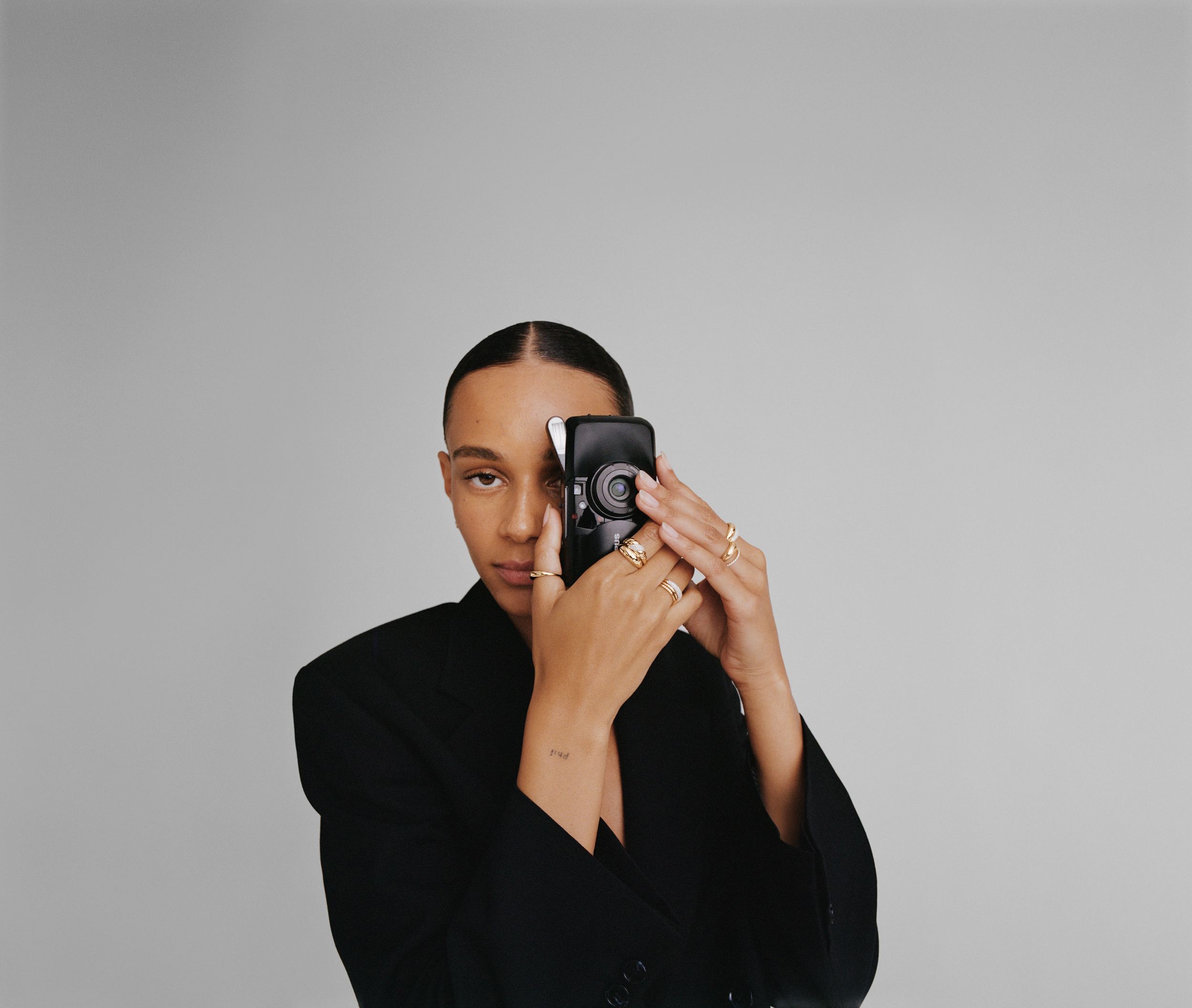 A woman with dark hair styled in a sleek bun, wearing a black blazer, is holding and looking through a small vintage camera with one eye, against a plain gray background.