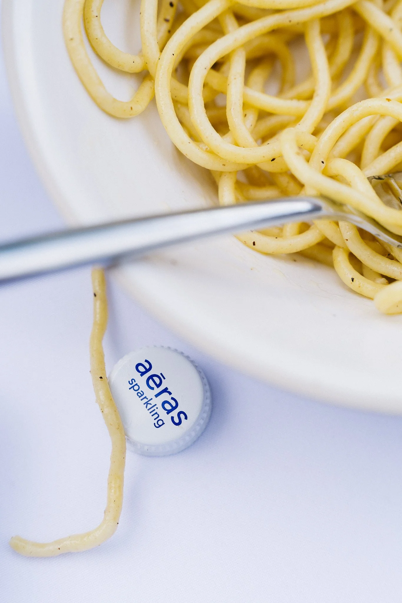 Close-up of a white bowl filled with cooked spaghetti, with a metal fork resting on the bowl's edge, and a bottle cap labeled 'aerates sparkling' on a white surface.