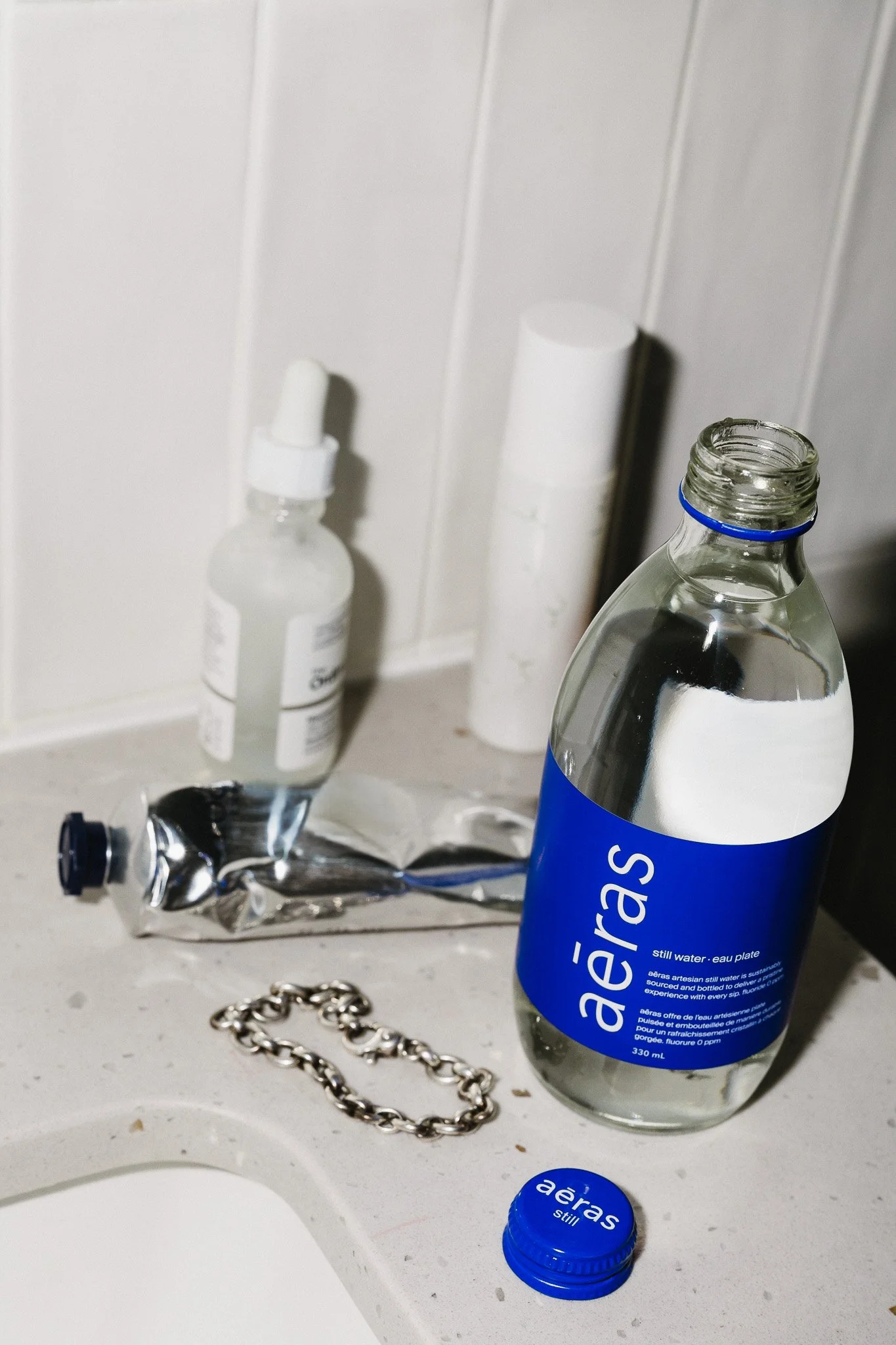 A bathroom countertop with a large plastic bottle of Aeras still water, a small dropper bottle, a cylindrical container, a crumpled metallic pouch, a chain bracelet, and a small blue bottle cap.