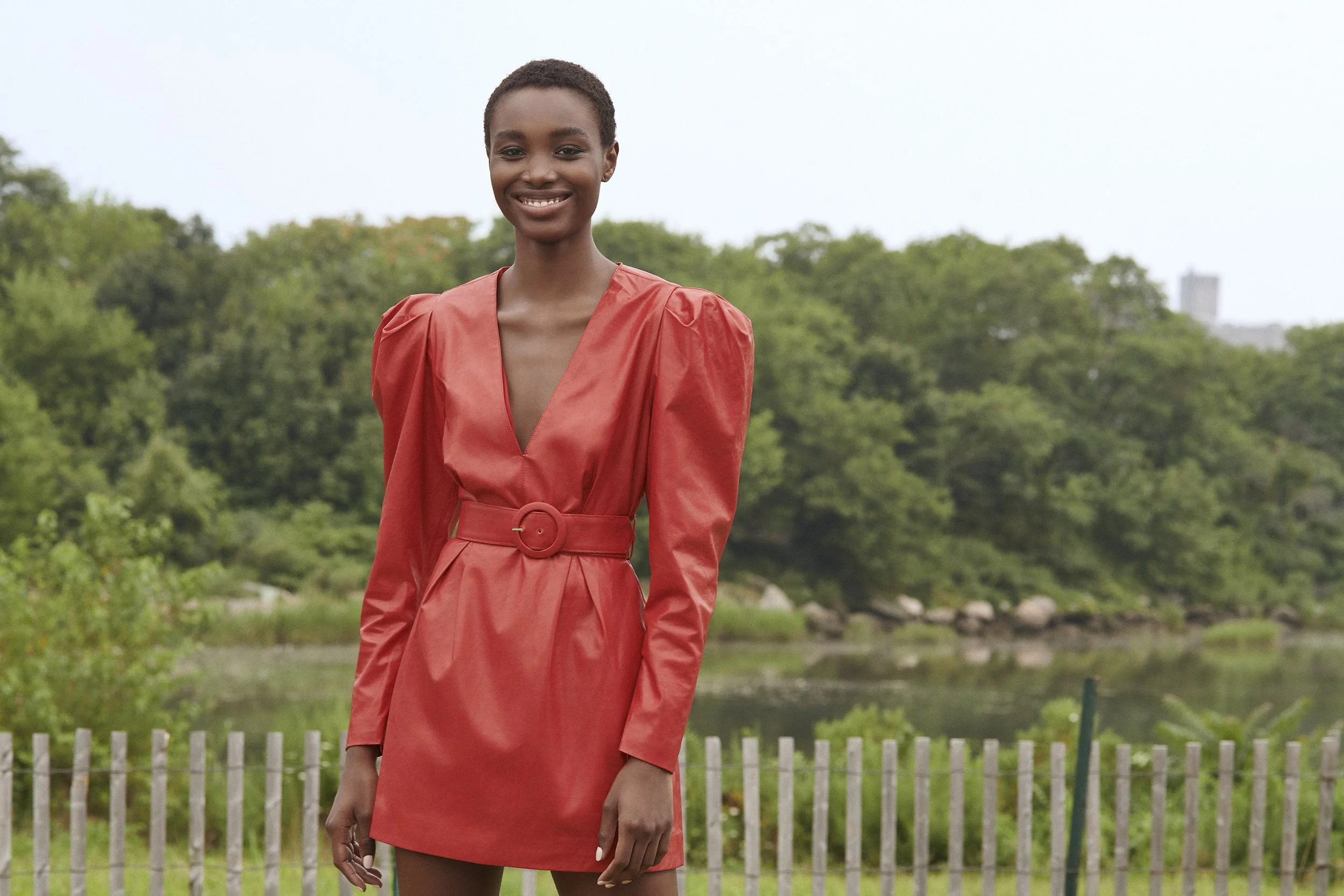 Young woman with short hair wearing a red dress with puffed sleeves, standing outdoors in front of greenery and a river, smiling at the camera.