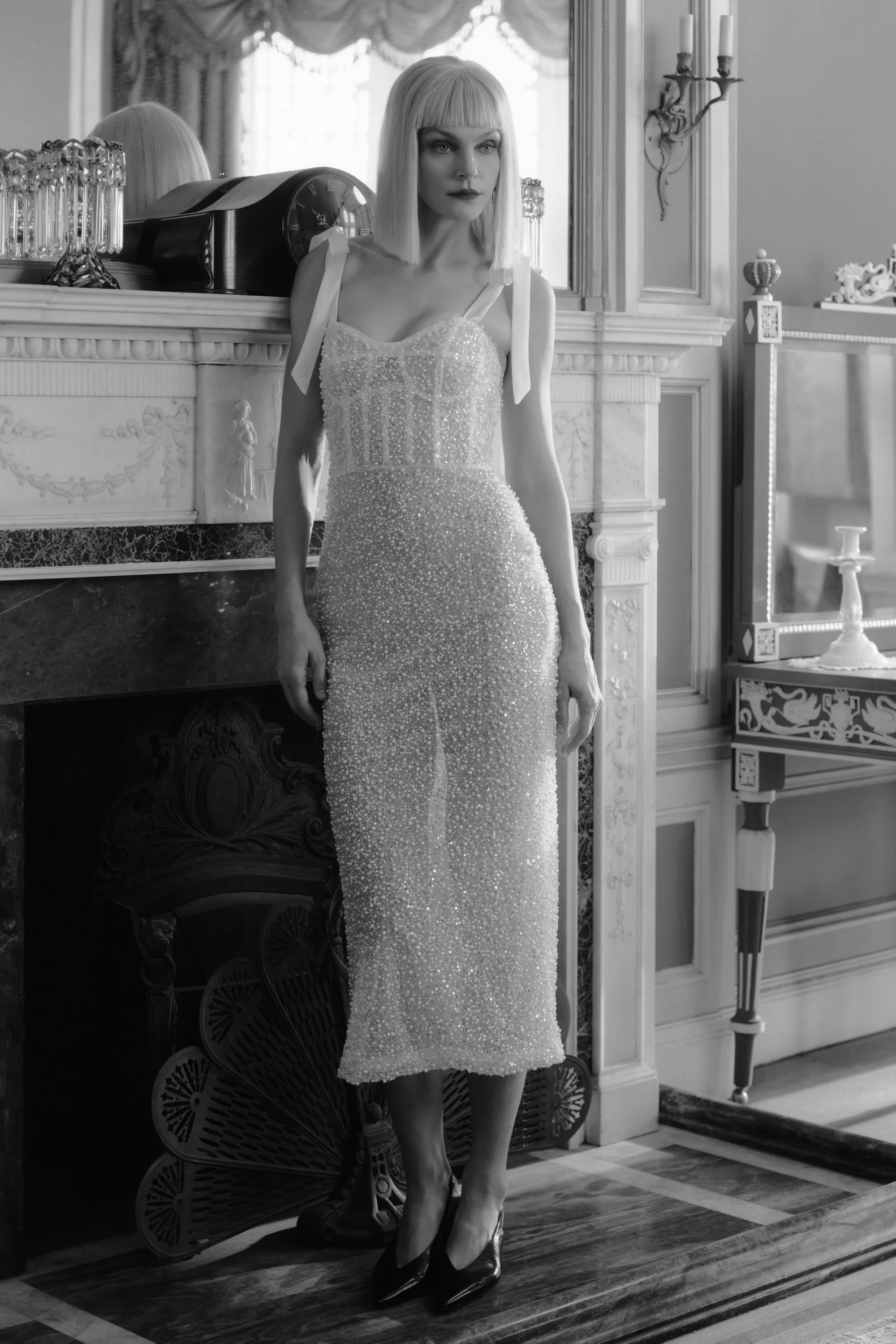 A woman in a sparkling, elegant dress standing in front of a fireplace in an ornate room, black and white photo.