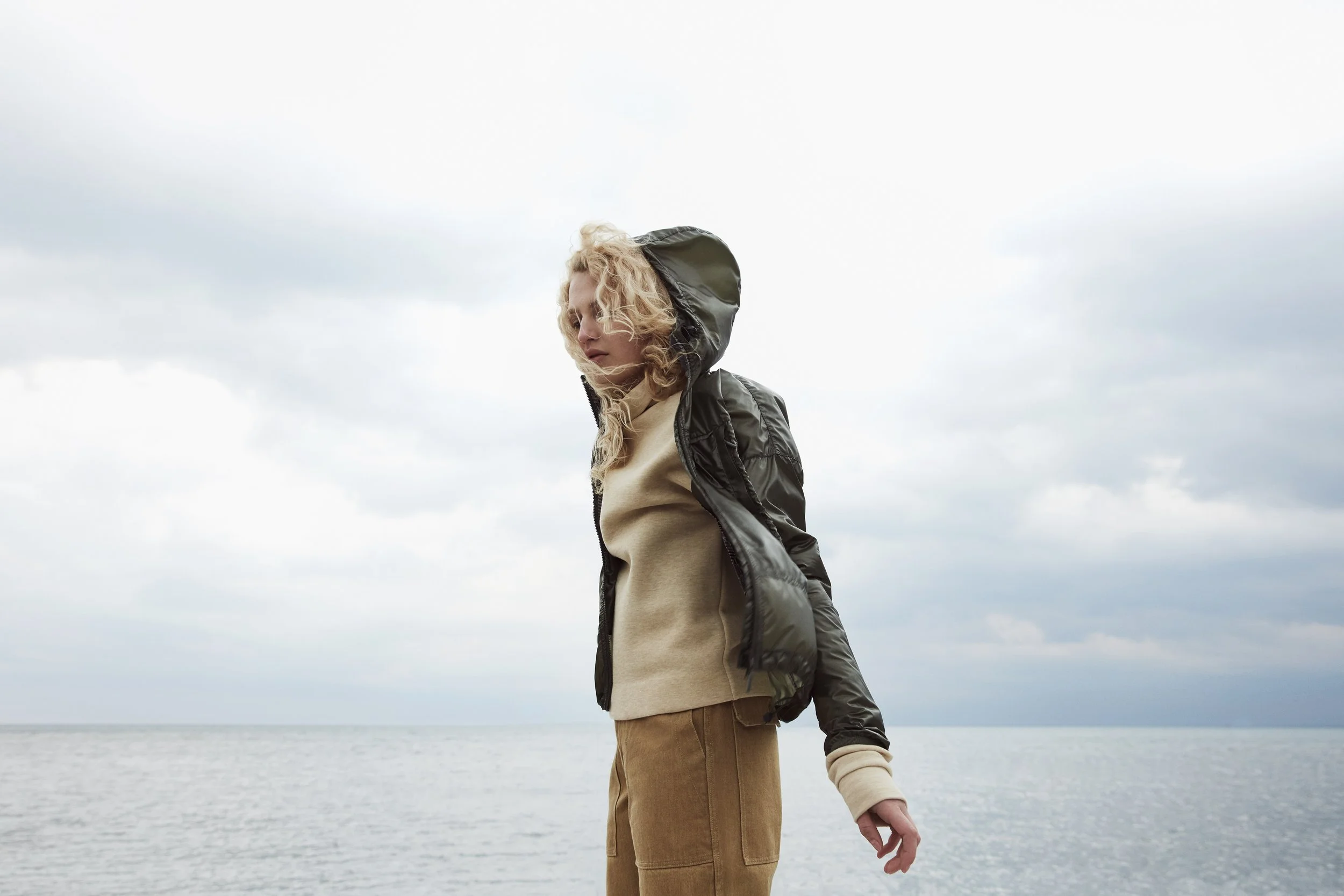 A woman standing outdoors by a body of water, wearing a beige sweater, brown pants, and a black jacket with the hood up, looking down with her eyes closed.