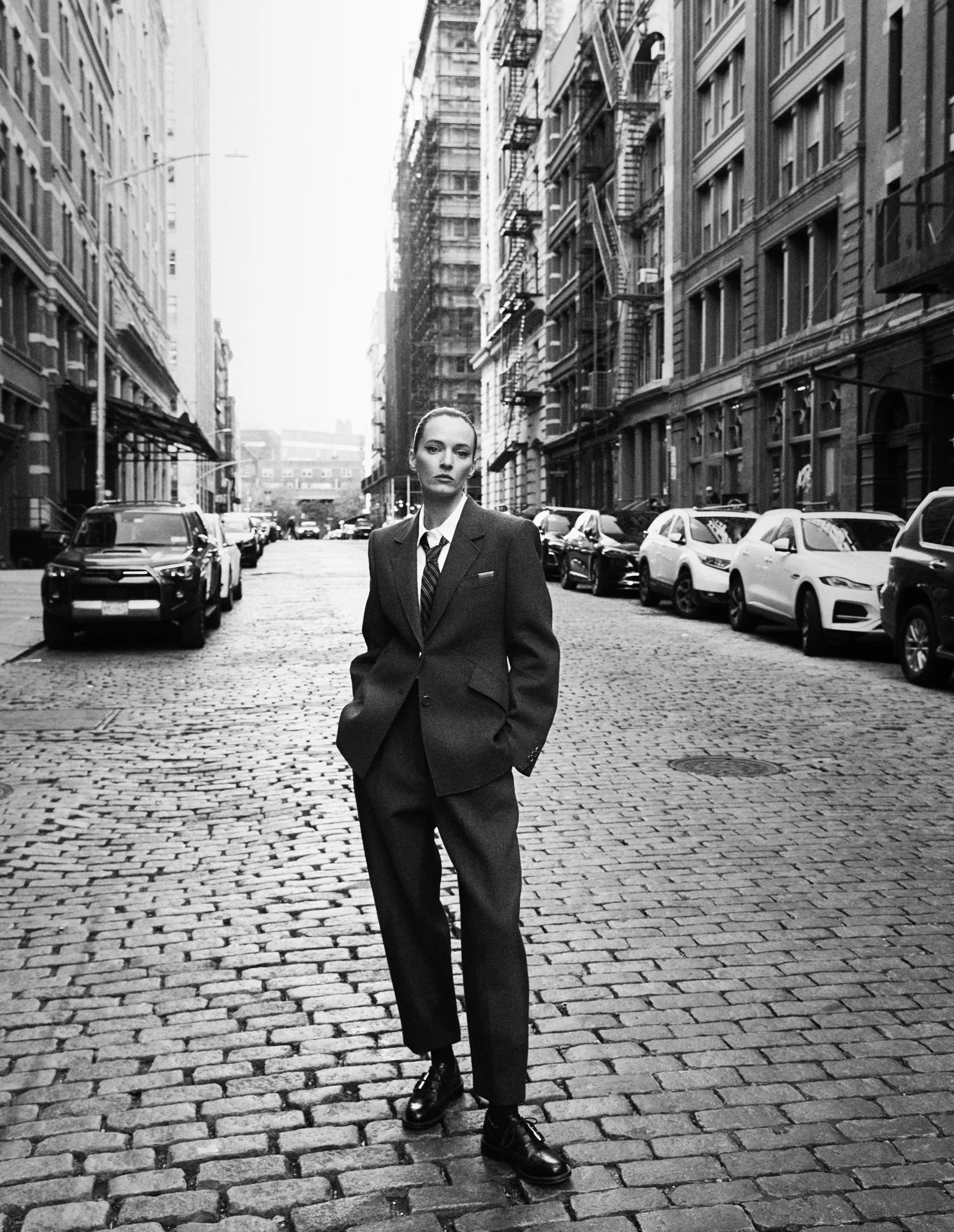 Black and white photo of a woman in a suit standing in the middle of a cobblestone street in an urban area with tall buildings and parked cars.