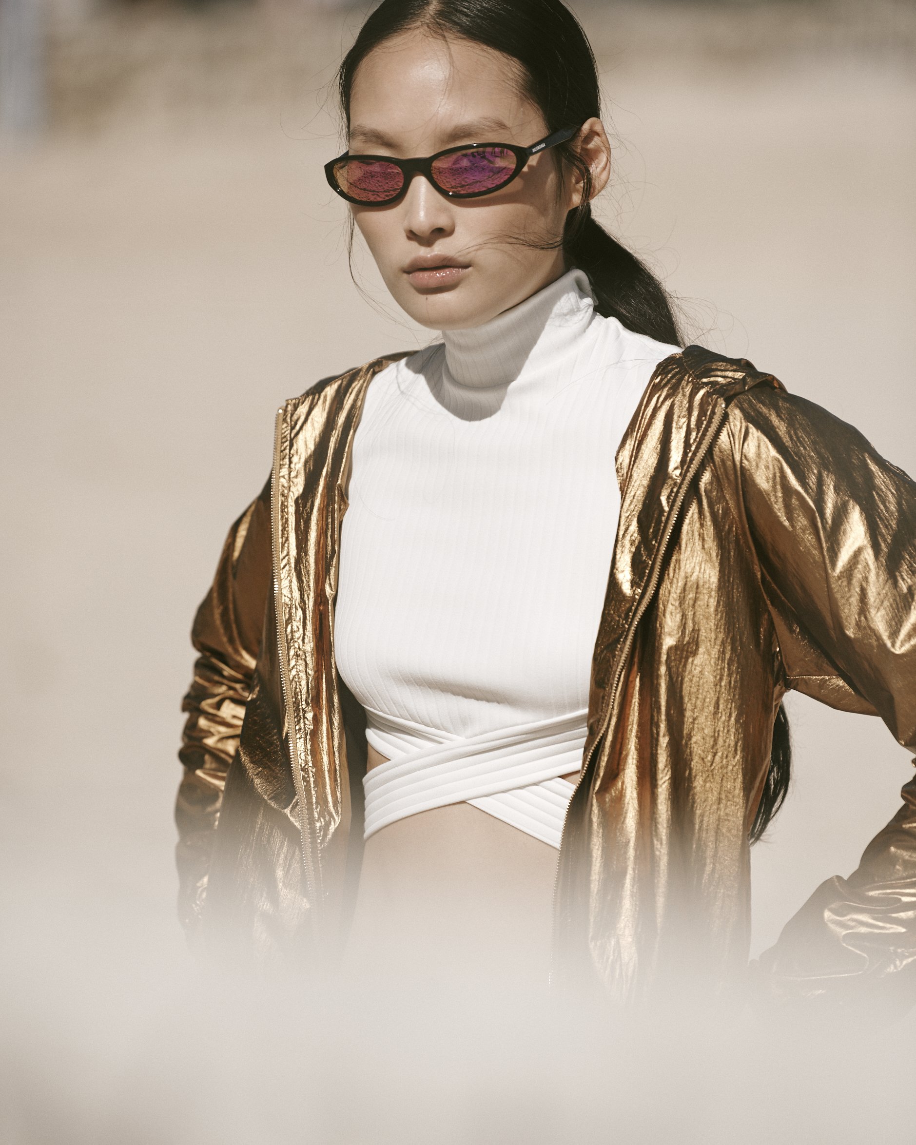 A woman wearing reflective sunglasses, a gold metallic jacket, and a white turtleneck top outdoors.