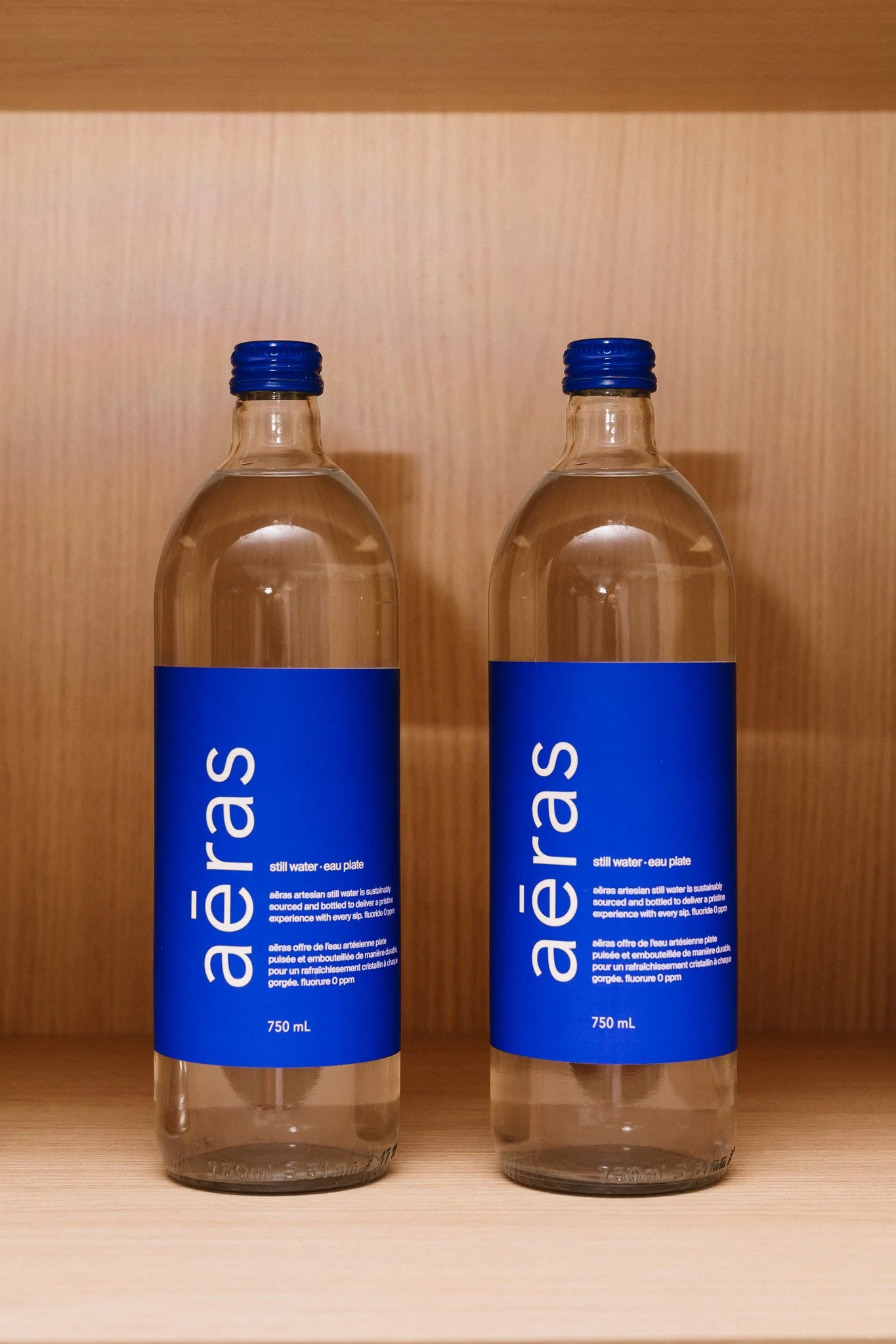 Two bottles of Aéras still water with blue labels and blue caps on a wooden surface against a wooden wall.