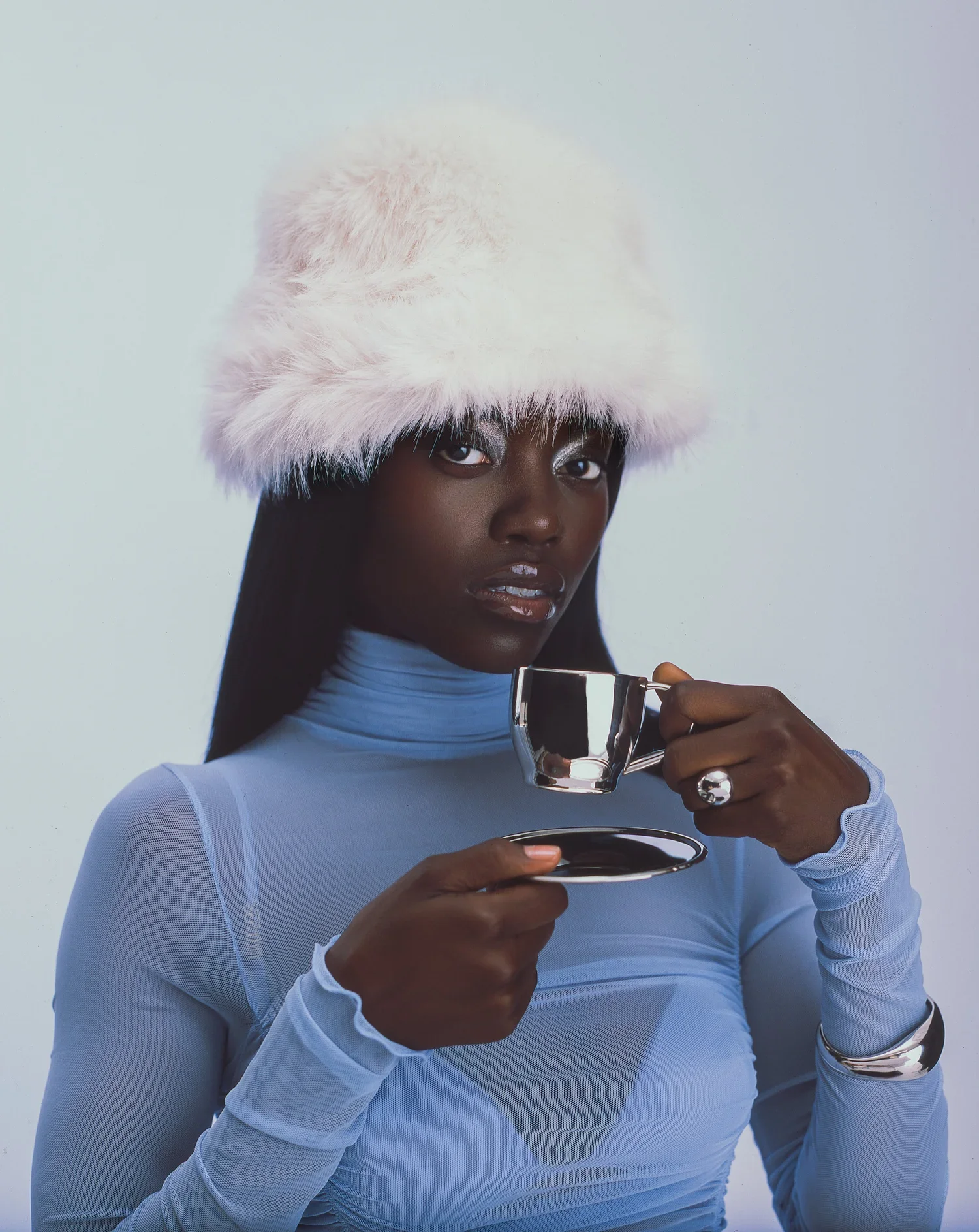 A woman with dark skin wearing a white fur hat and a light blue sheer top holding a silver teacup and saucer, with a neutral background.